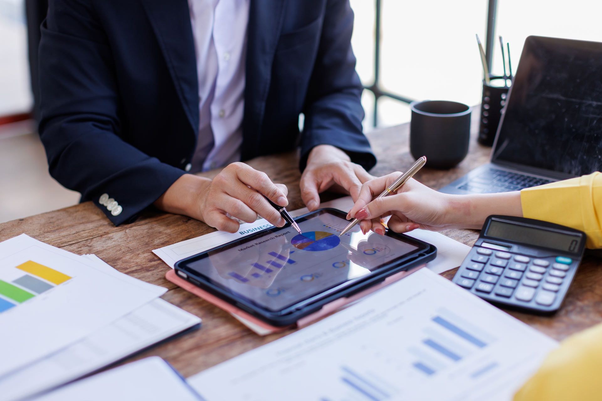 Two people analyzing charts on a tablet, with laptop, calculator, and papers on a desk.