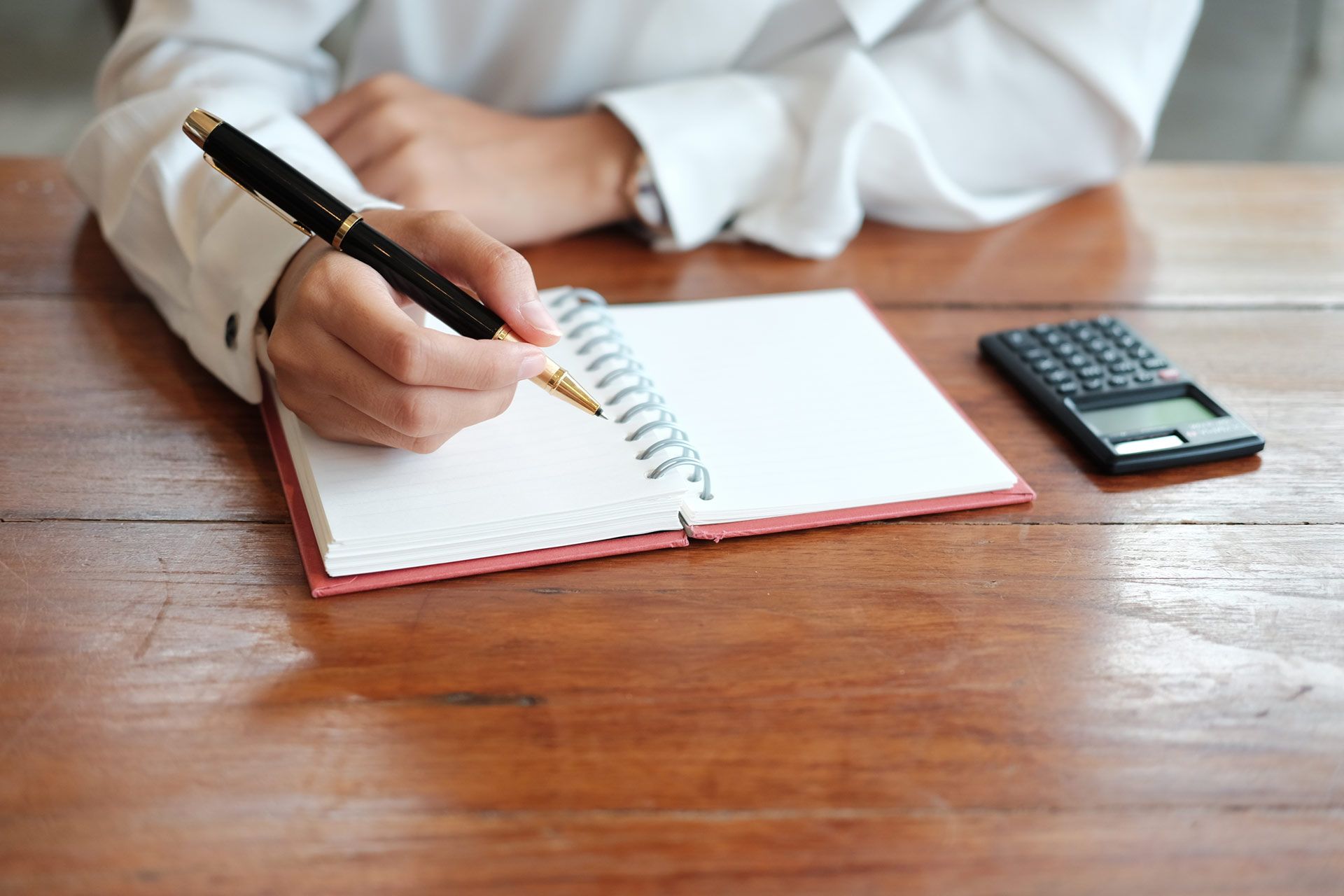 Person writing in a notebook with a pen at a desk, a calculator nearby.