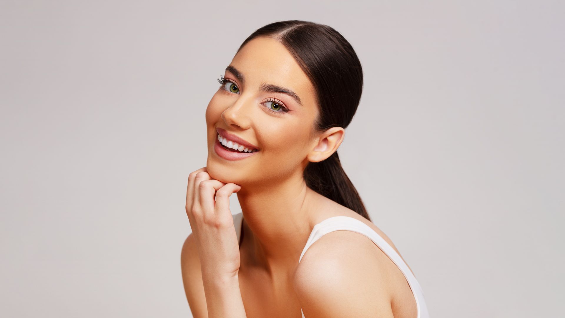 A woman in a white tank top is smiling and touching her face.