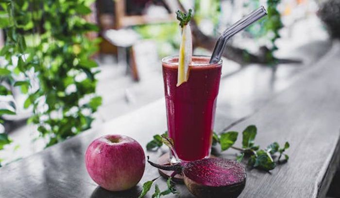 A glass of red juice with a straw and an apple on a wooden table.