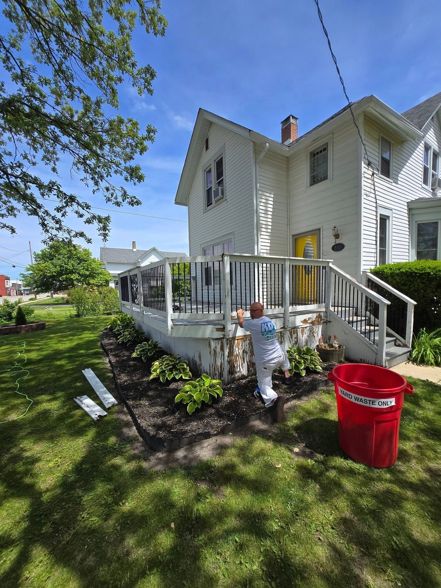 A child is standing in front of a white house next to a red bucket.