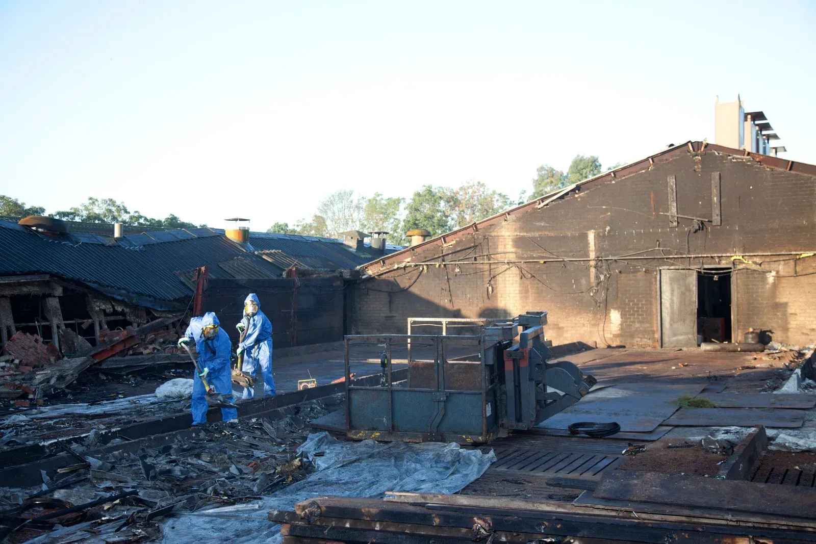 Two figures in blue protective suits survey fire damage on a rooftop. Burned buildings, debris.