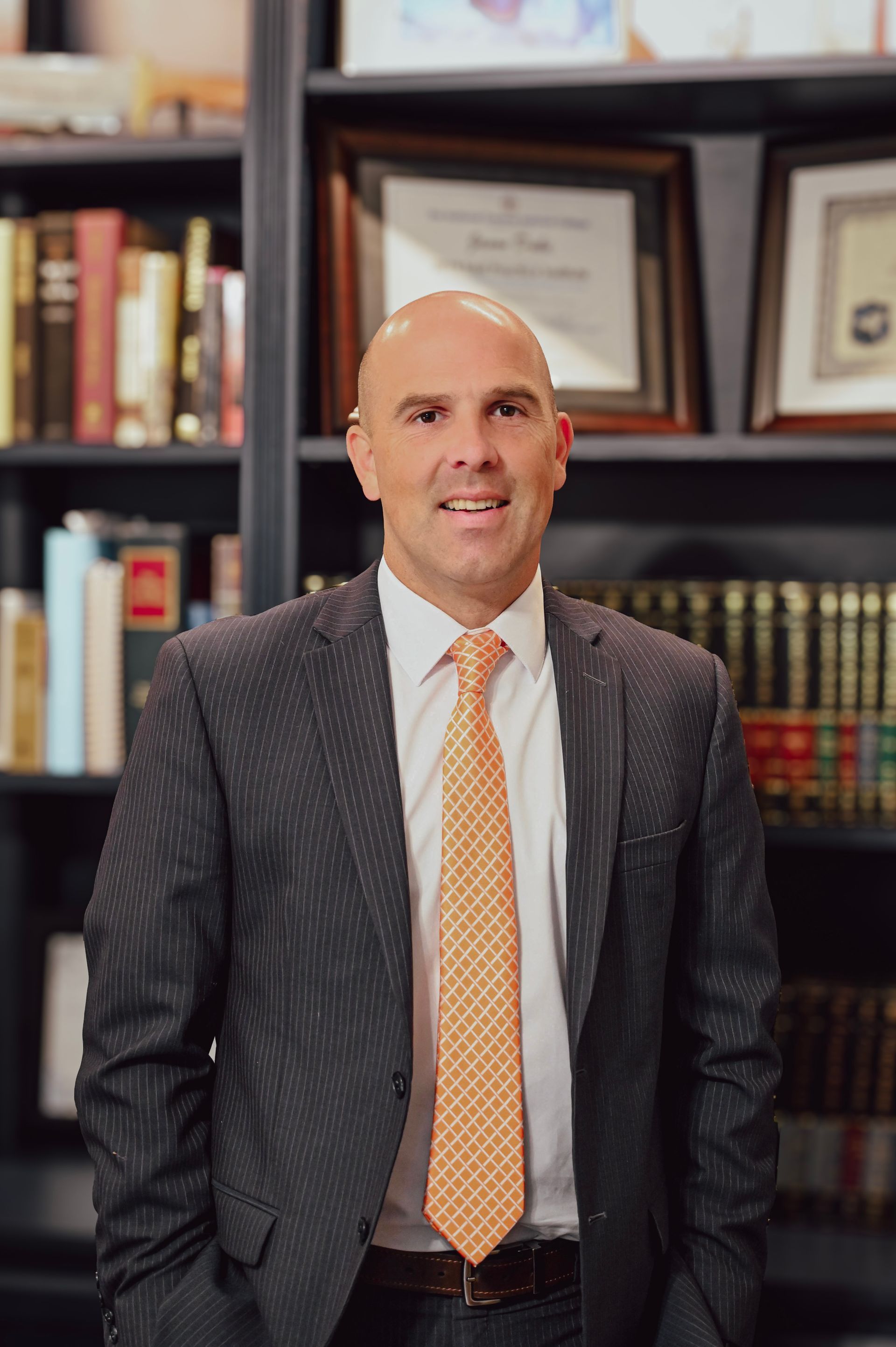 Business professional wearing a dark pinstripe suit and orange patterned tie, standing in an office with bookshelves displaying law books and framed certificates in the background