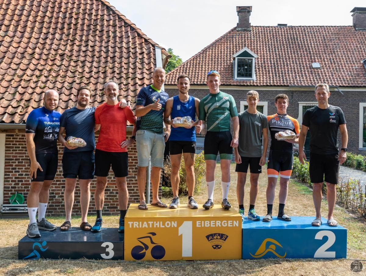Triathlon Eibergen. Een groep mannen staat op een podium voor een bakstenen gebouw.