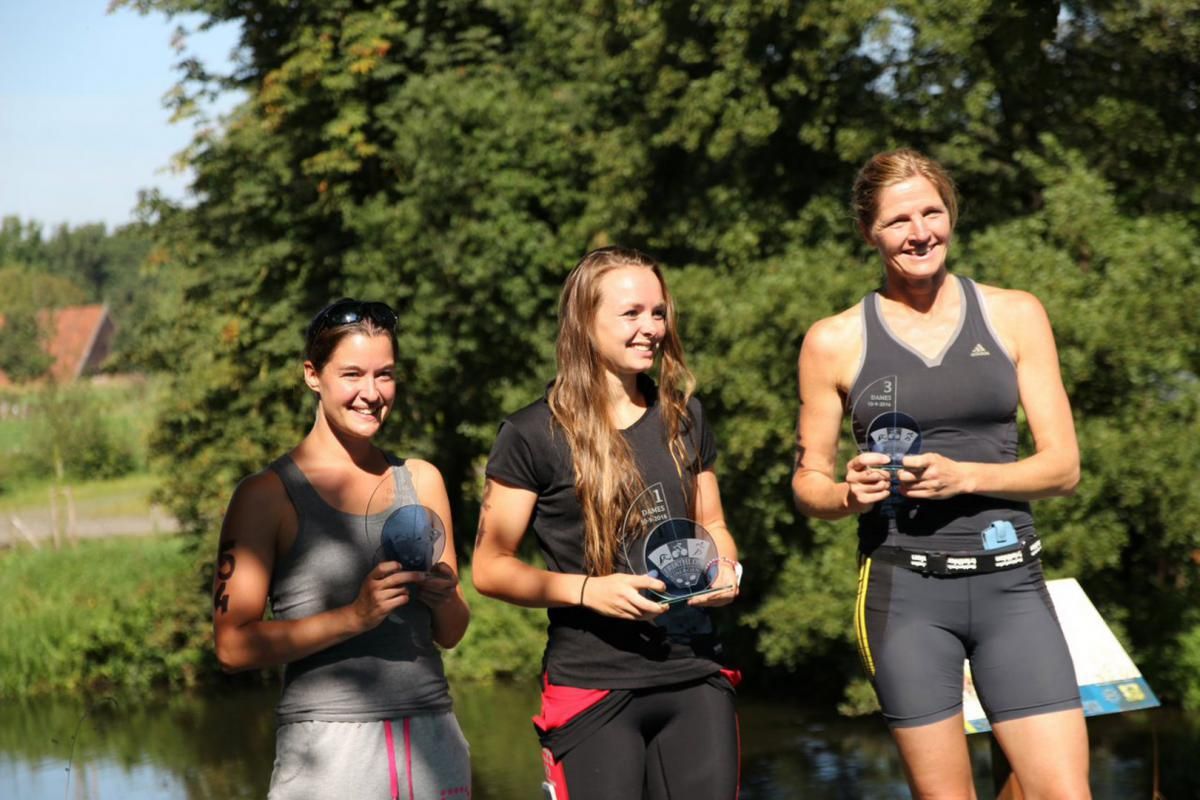 Drie vrouwen staan ​​naast elkaar voor een waterpartij en houden trofeeën vast.