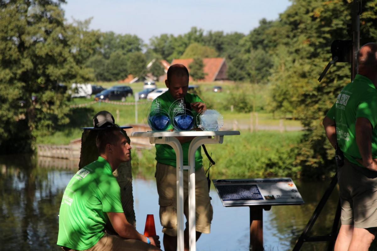 Een groep mensen in groene shirts staat rond een tafel voor een meer.