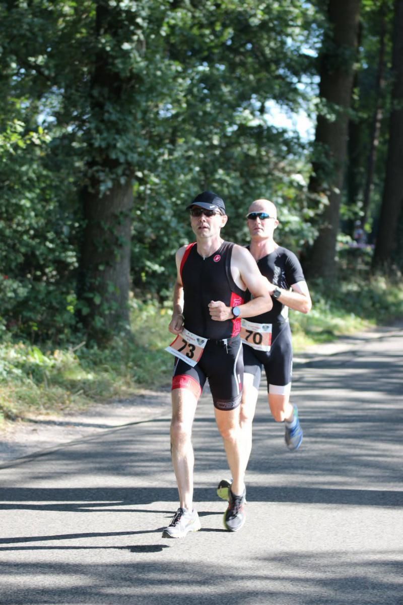 Twee mannen rennen over een weg met bomen op de achtergrond.