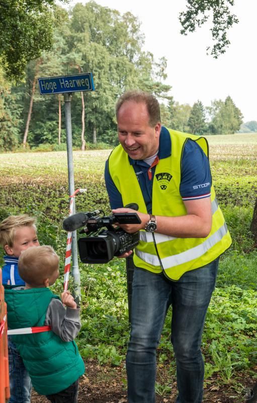Een man in een geel vest houdt een camera vast terwijl twee kinderen toekijken.