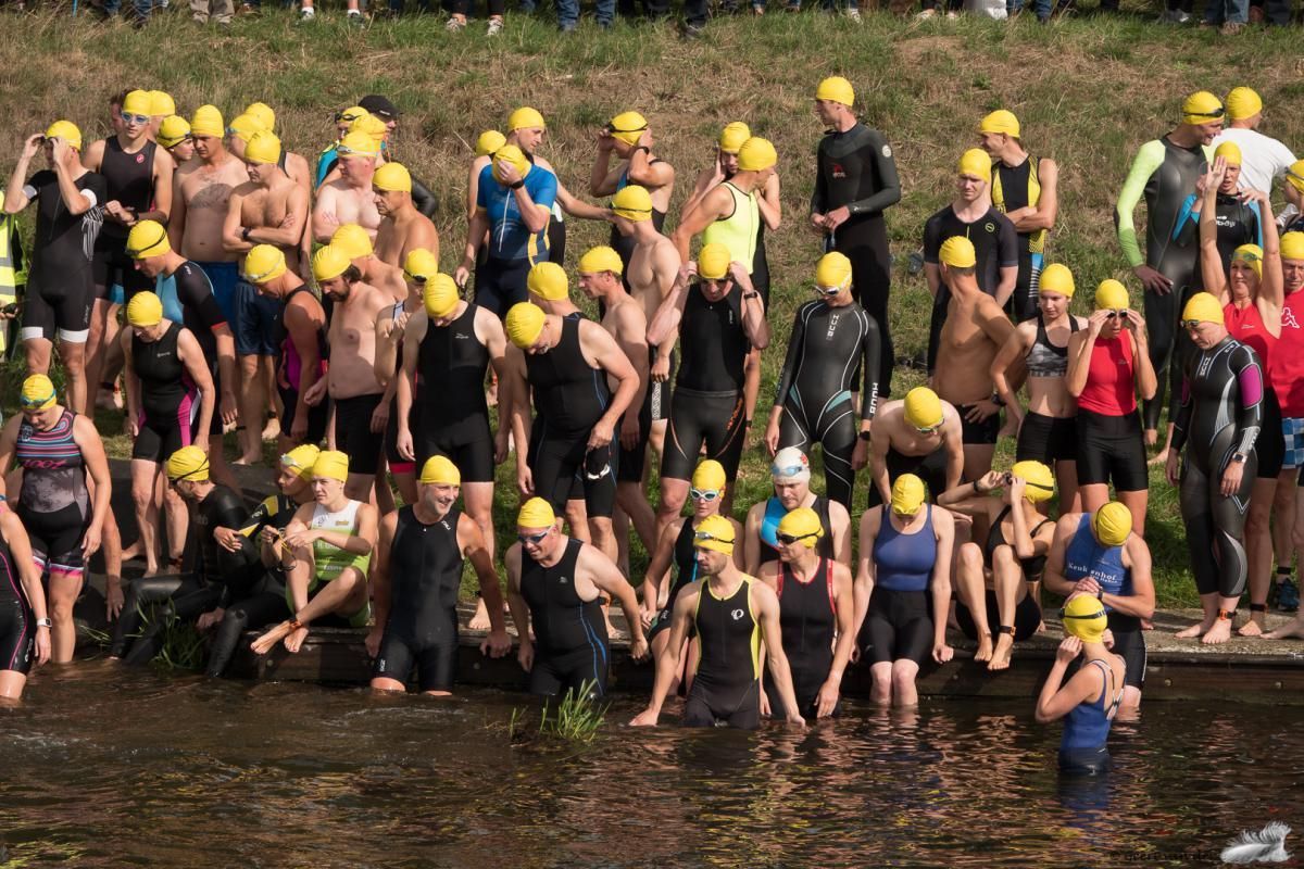 Een grote groep mensen met wetsuits en gele badmutsen staat in het water.