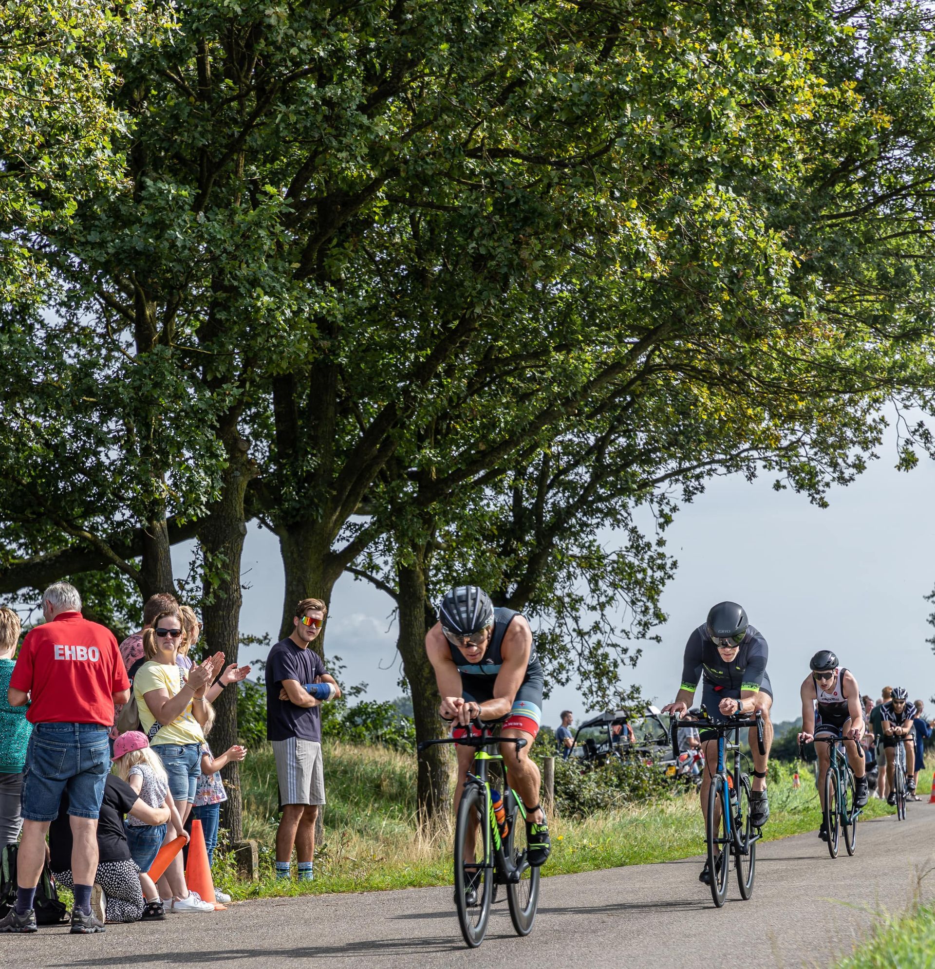 Een groep fietsers rijdt over een weg met een man die een rood shirt draagt ​​met de tekst chris