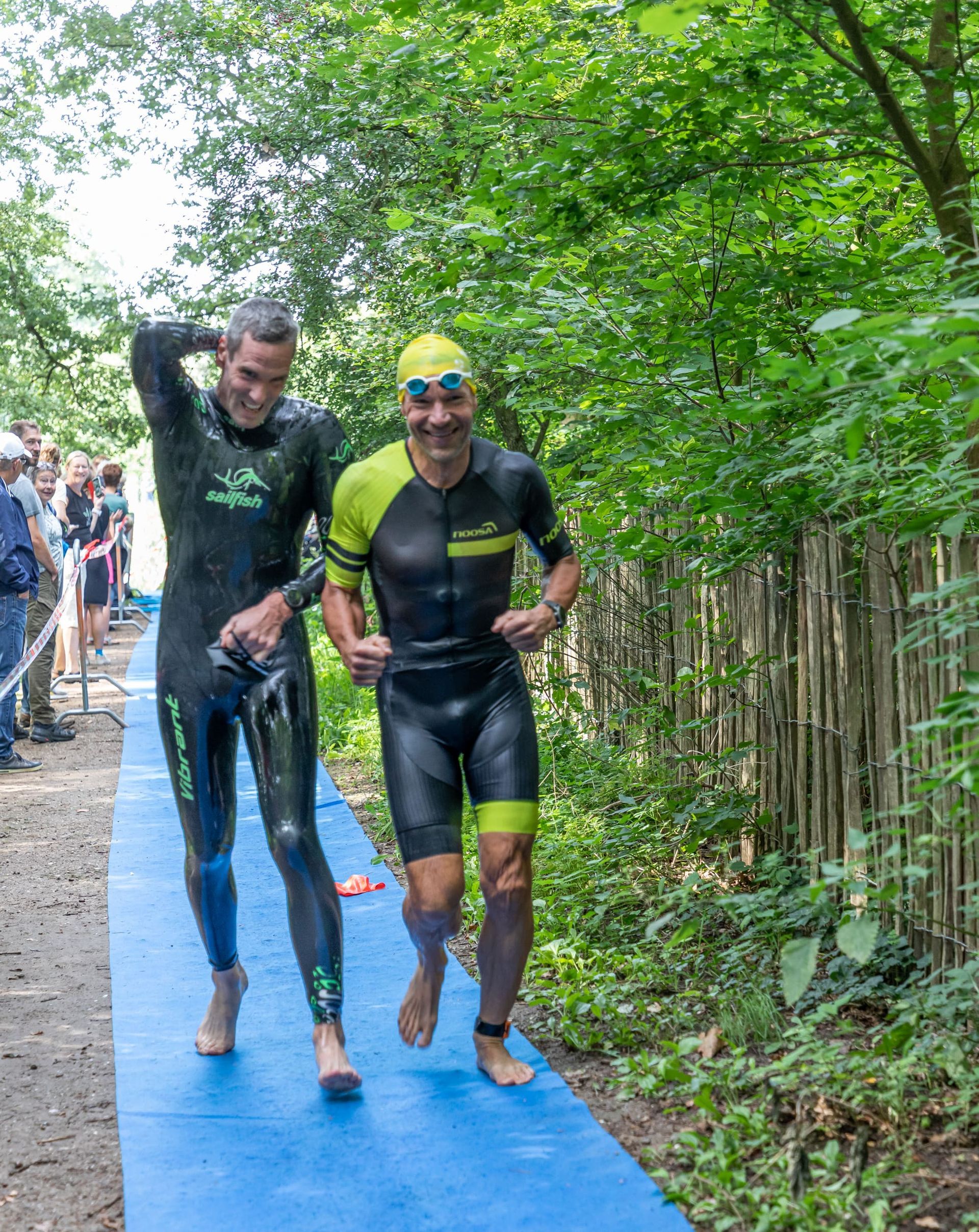 Twee mannen in wetsuits lopen over een pad in het bos.
