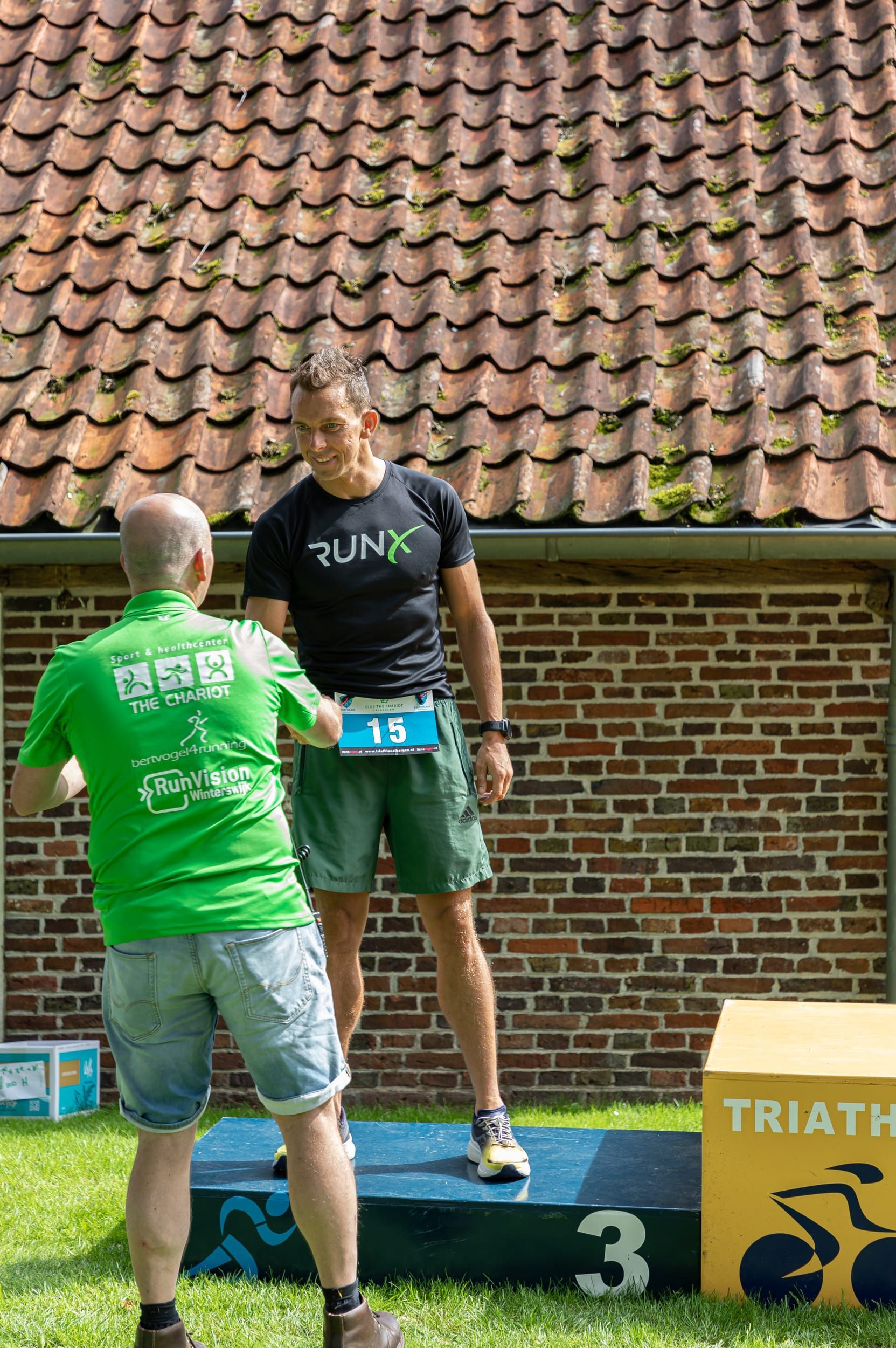 Prijsuitreiking Triathlon Eibergen 2023. Twee mannen staan ​​op een podium voor een bakstenen gebouw.