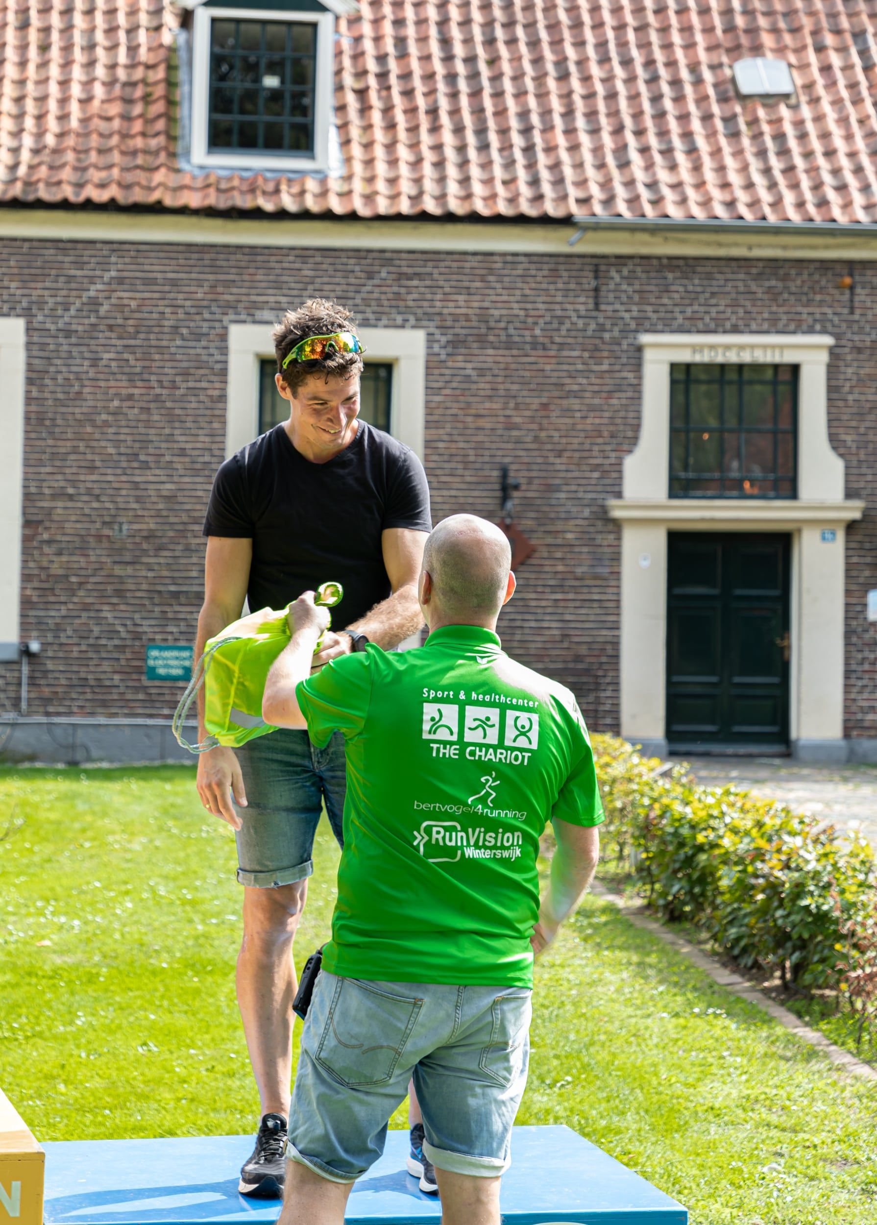 Prijsuitreiking Triathlon Eibergen 2023. Een man in een groen shirt geeft een geschenk aan een andere man voor een bakstenen gebouw.