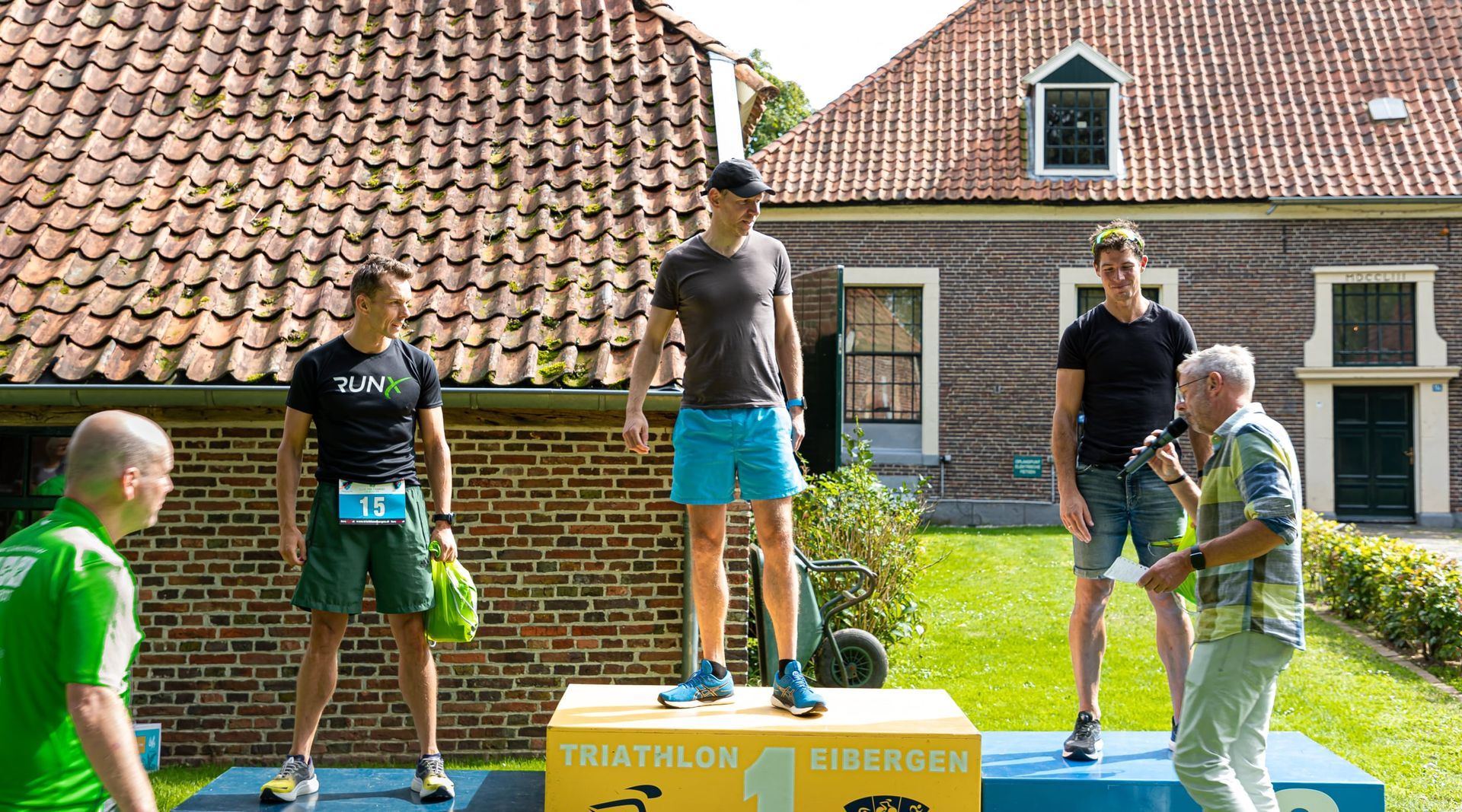 Prijsuitreiking Triathlon Eibergen 2023. Een groep mannen staat op een podium voor een bakstenen gebouw.