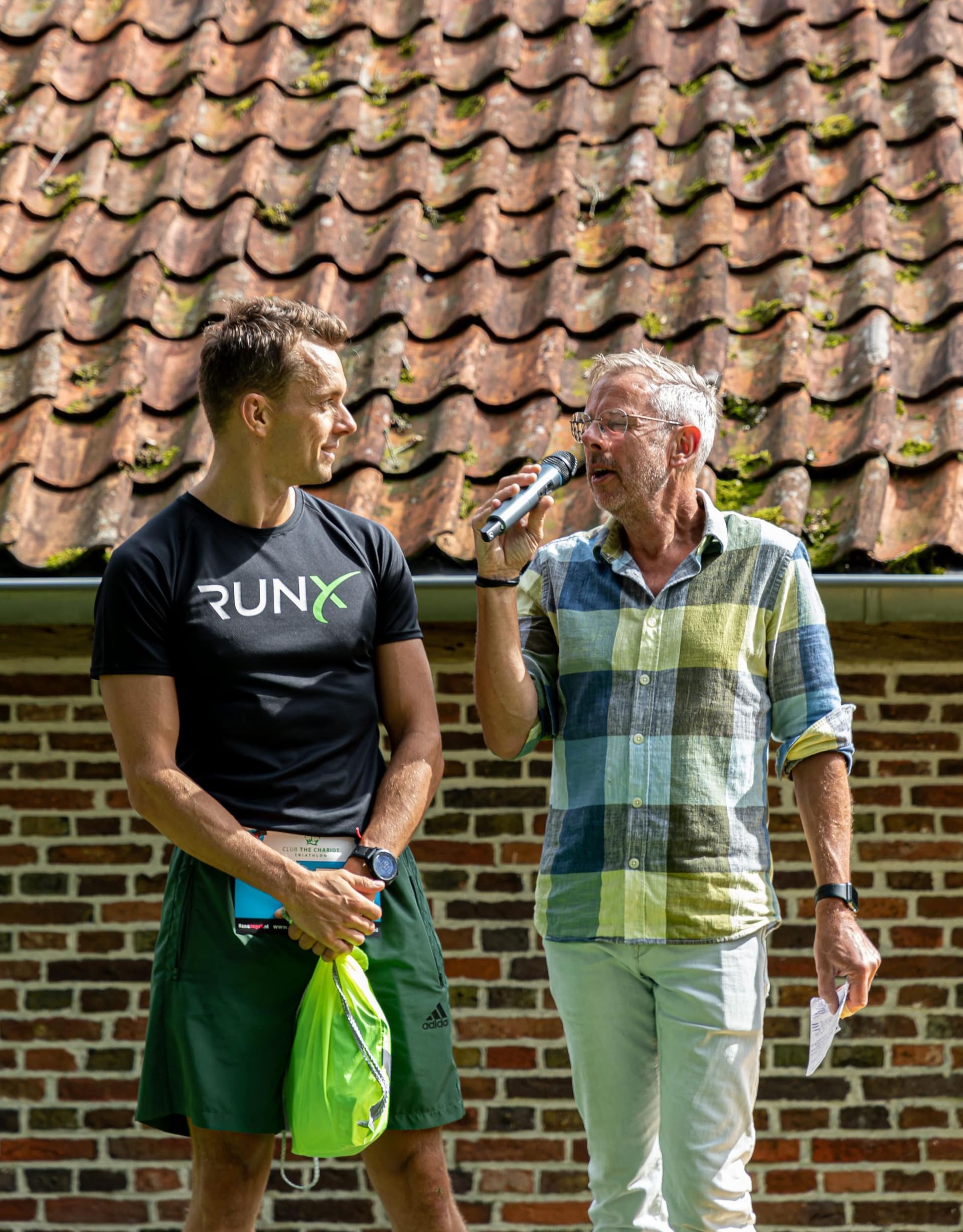 Prijsuitreiking Triathlon Eibergen 2023. Twee mannen staan ​​naast elkaar en praten via microfoons voor een bakstenen gebouw.