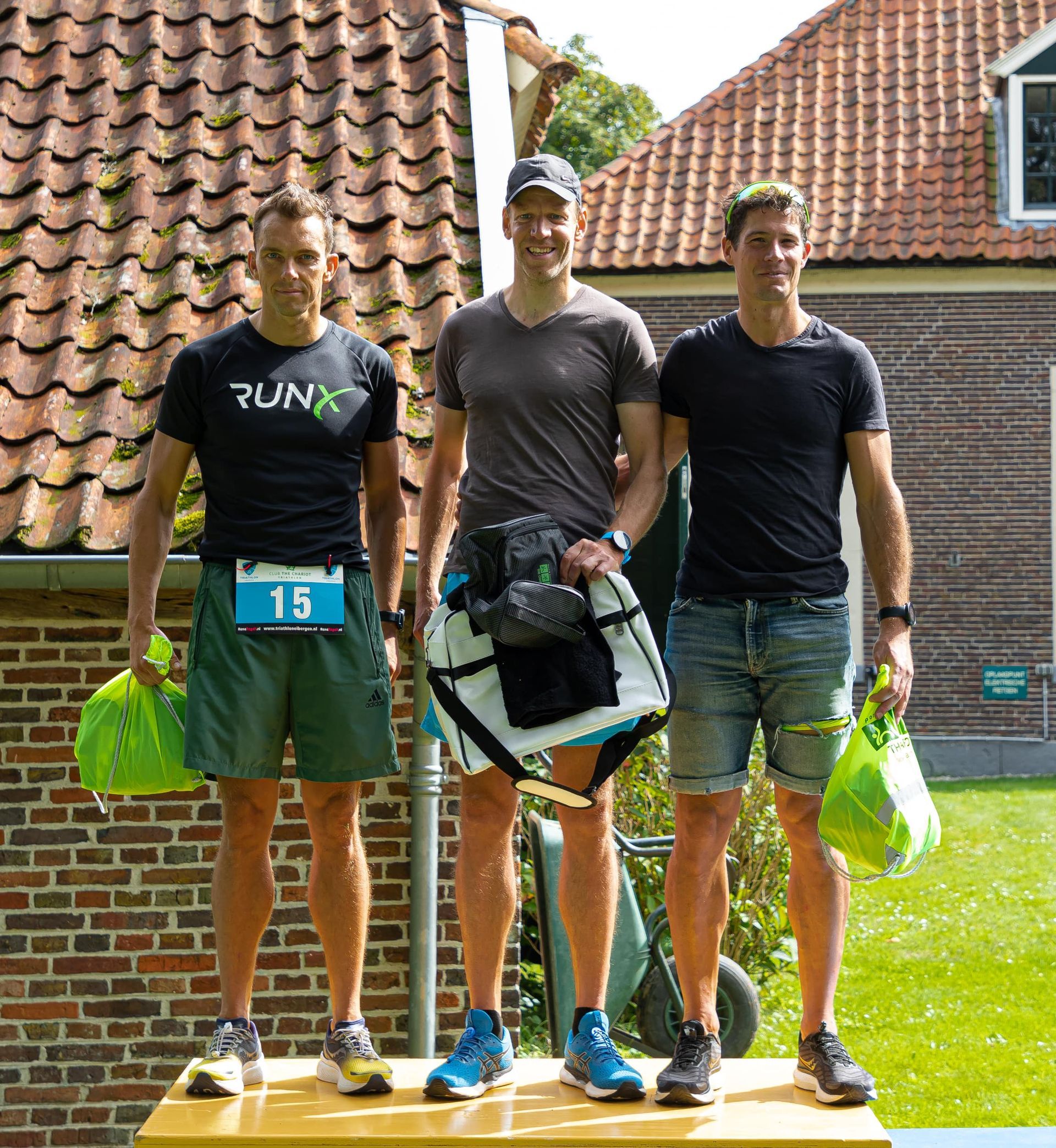 Prijsuitreiking Triathlon Eibergen 2023. Drie mannen staan ​​op een podium voor een bakstenen gebouw met tassen in hun handen.