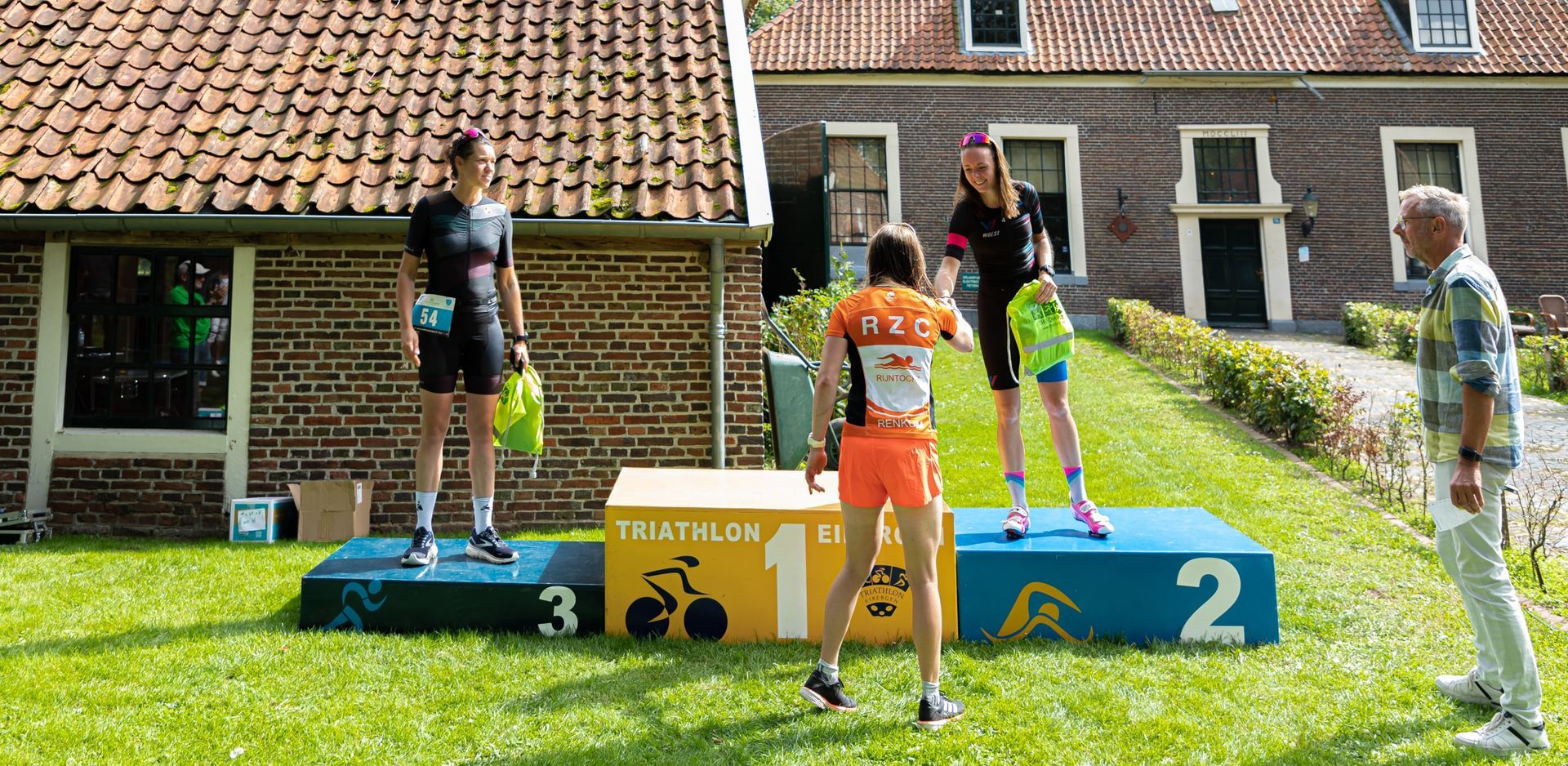 Prijsuitreiking Triathlon Eibergen 2023. Een groep mensen staat op een podium voor een bakstenen gebouw.