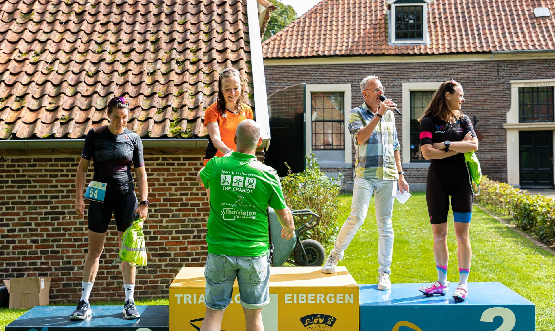 Prijsuitreiking Triathlon Eibergen 2023. Een groep mensen die op een podium voor een bakstenen gebouw staat.