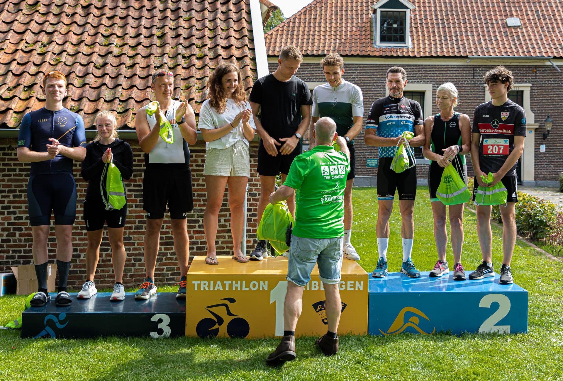 Prijsuitreiking Triathlon Eibergen 2023. Een groep mensen staat op een podium voor een bakstenen gebouw.