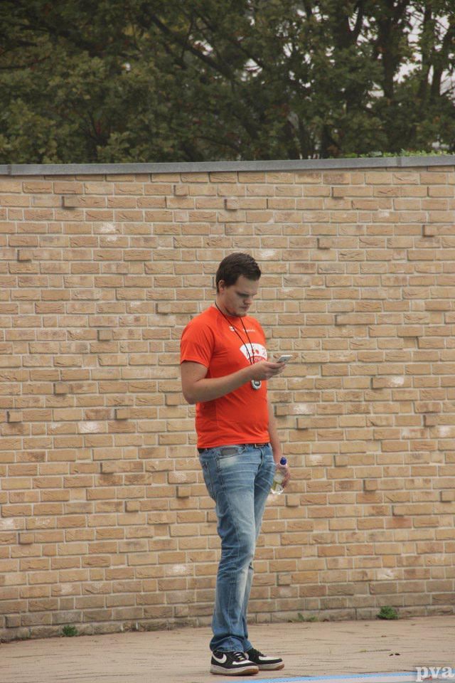 Triathlon Eibergen. Een man in een rood shirt staat voor een bakstenen muur en kijkt op zijn telefoon.