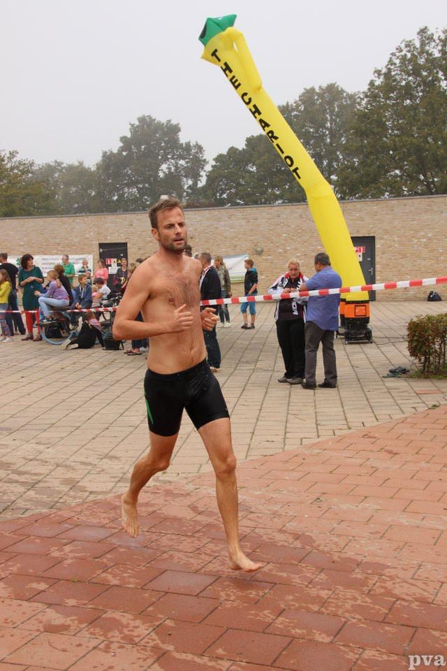 Triathlon Eibergen. Een man zonder shirt rent op blote voeten voor een geel opblaasbaar voorwerp met de tekst 'triatlon' erop.
