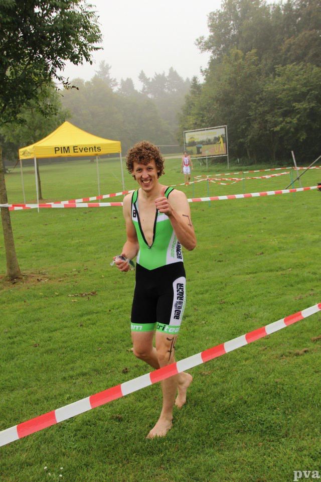 Triathlon Eibergen. Een man rent op blote voeten door een veld met een gele tent op de achtergrond.