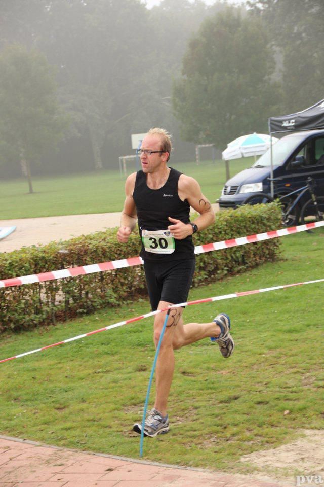 Triathlon Eibergen. Een man in een zwart tanktopje en korte broek rent over een grasveld.