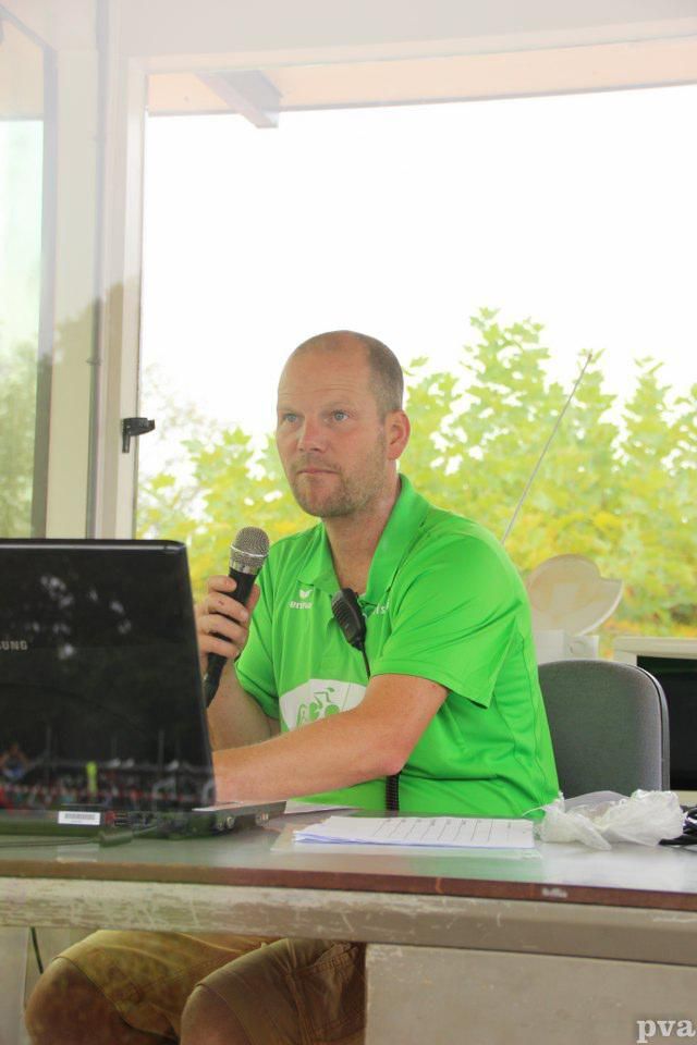 Een man in een groen shirt zit aan een bureau en houdt een microfoon vast.