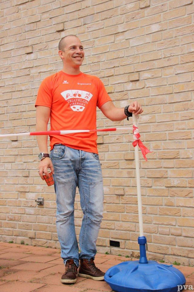 Een man in een oranje shirt staat voor een bakstenen muur met een hoelahoep in zijn handen.