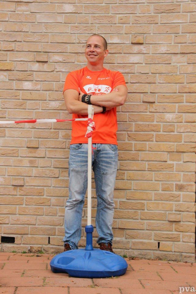 Een man in een oranje shirt staat voor een bakstenen muur met zijn armen over elkaar.