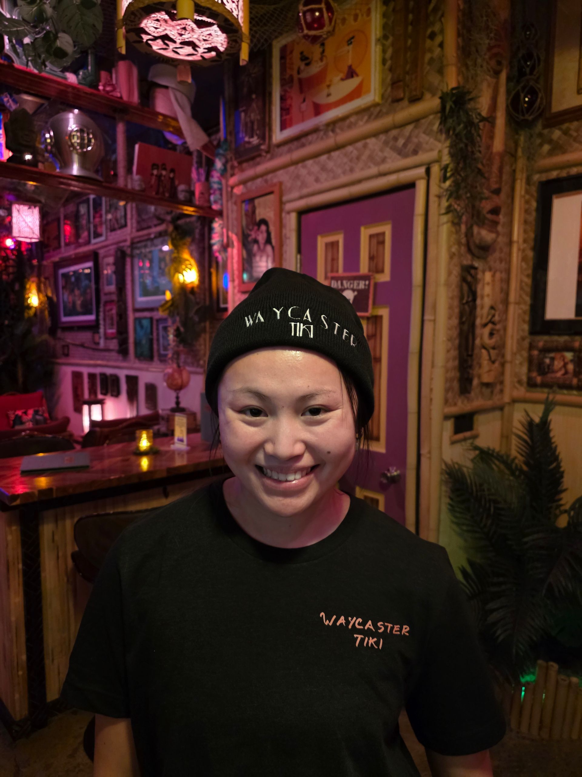 Person in a black beanie and shirt smiling in a tiki-themed bar, surrounded by decor.