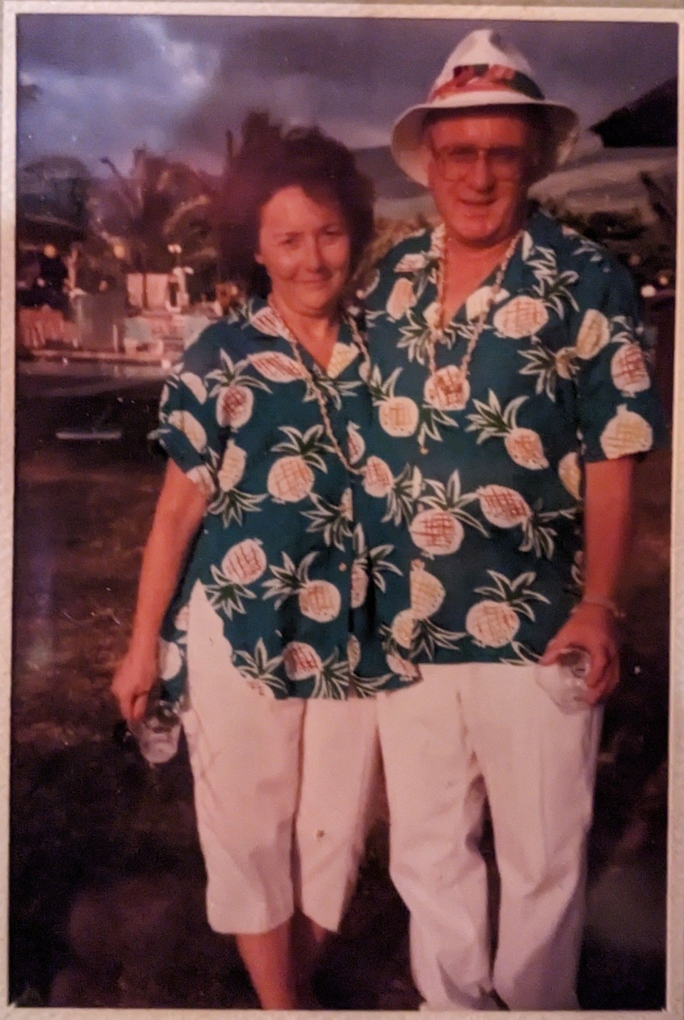 Couple in matching pineapple print shirts and white pants, posing outdoors.