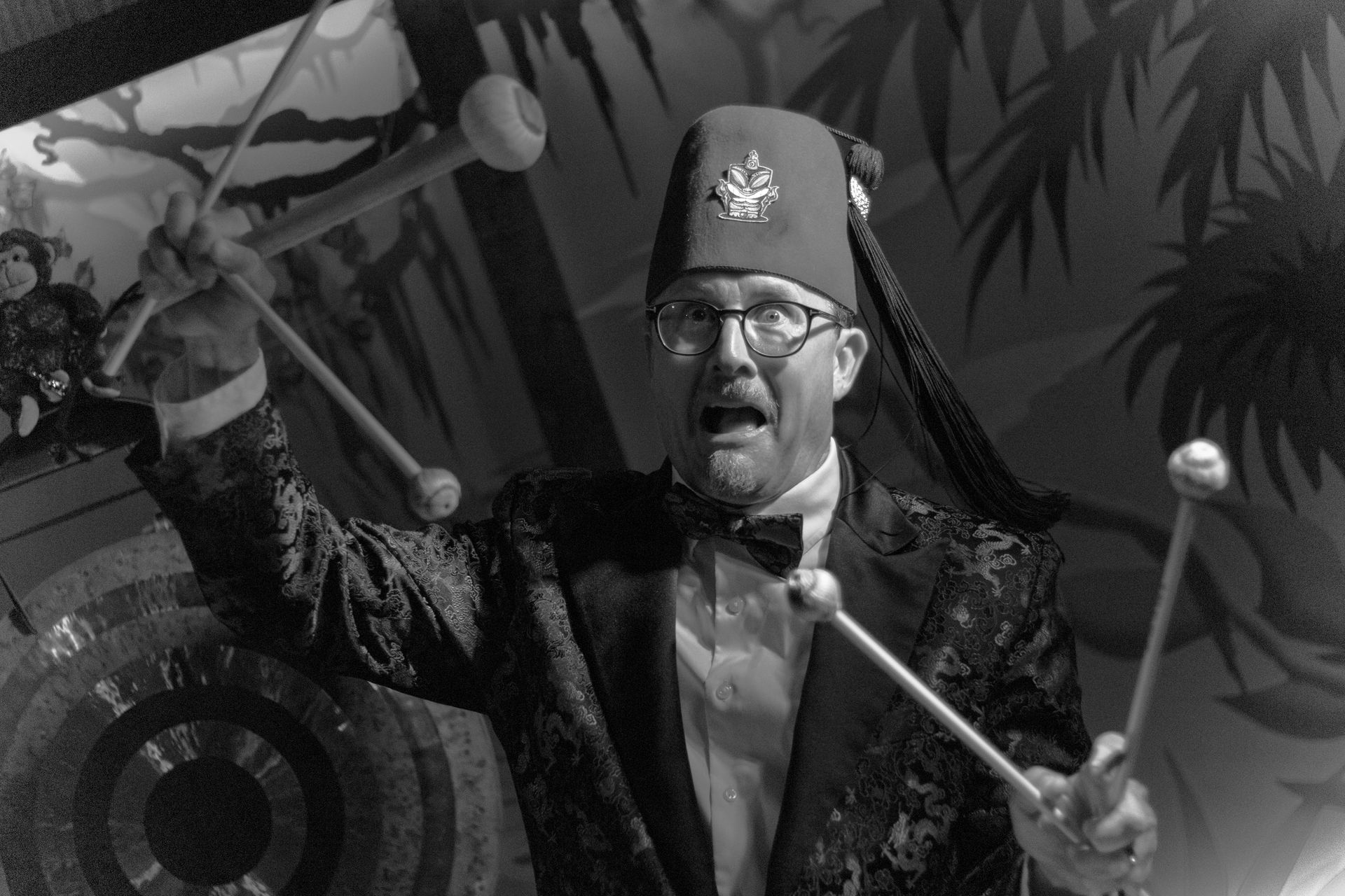 Man in fez and tuxedo strikes a gong, wide-eyed expression, indoor setting.