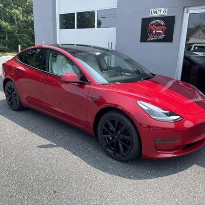 A red tesla model 3 is parked in front of a garage.