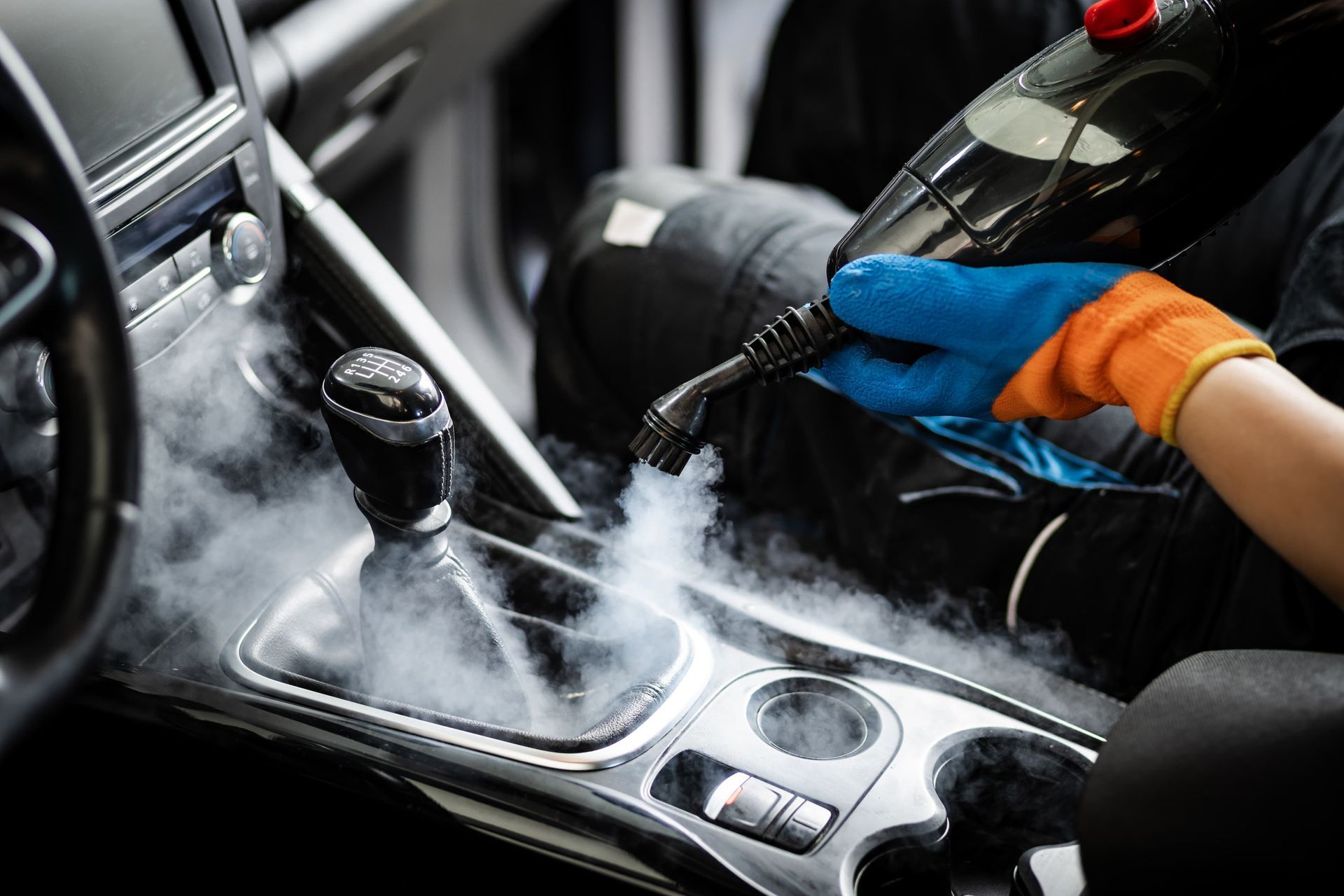 Person steam cleaning a car interior. Blue and orange gloves, steam, black car features.
