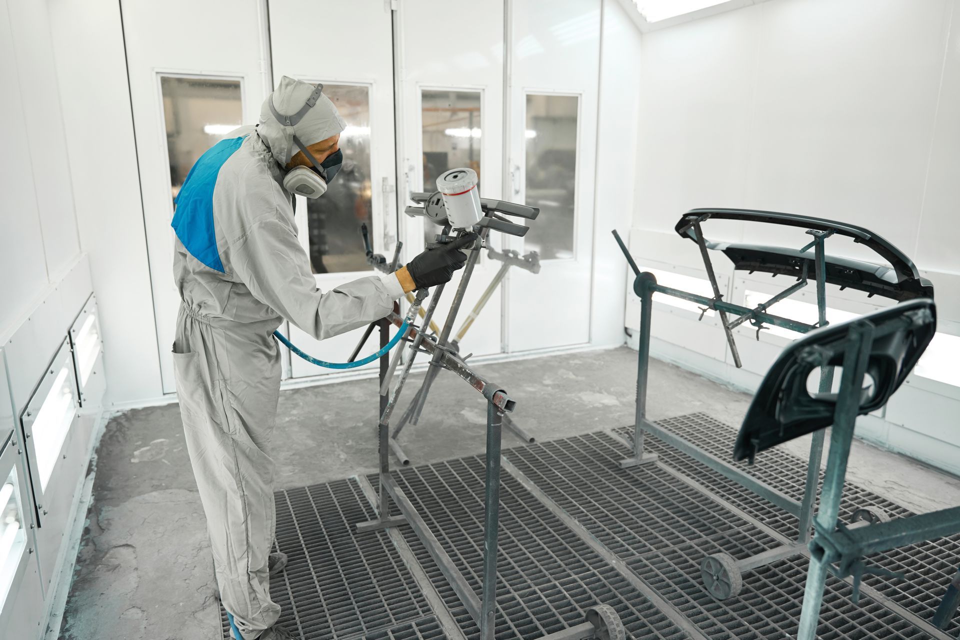 A person in a protective suit and respirator sprays paint onto an automotive part in a bright, white spray booth.