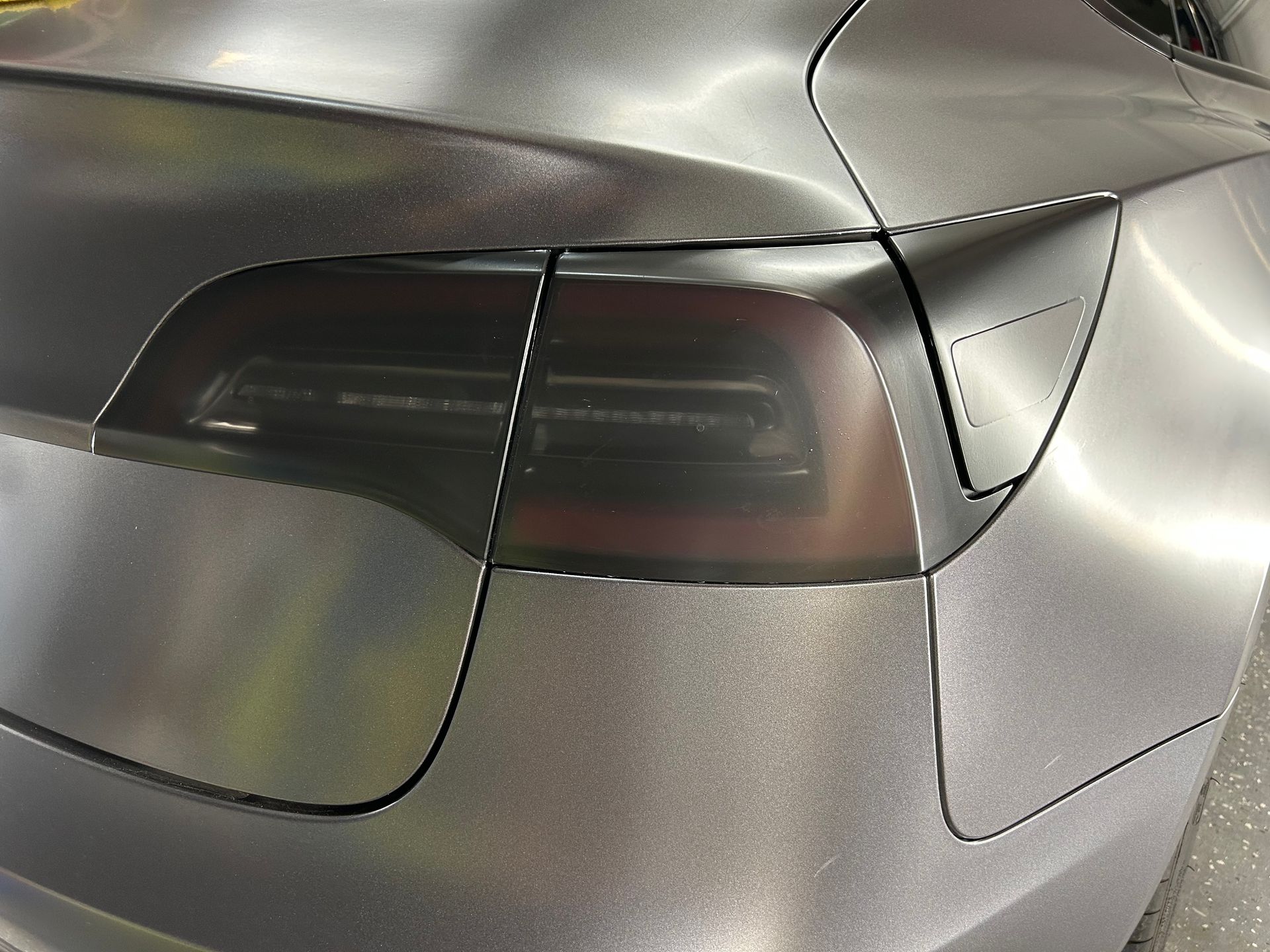 A matte gray Tesla Model 3 tail light with a dark tinted lens cover on a metallic gray car body.