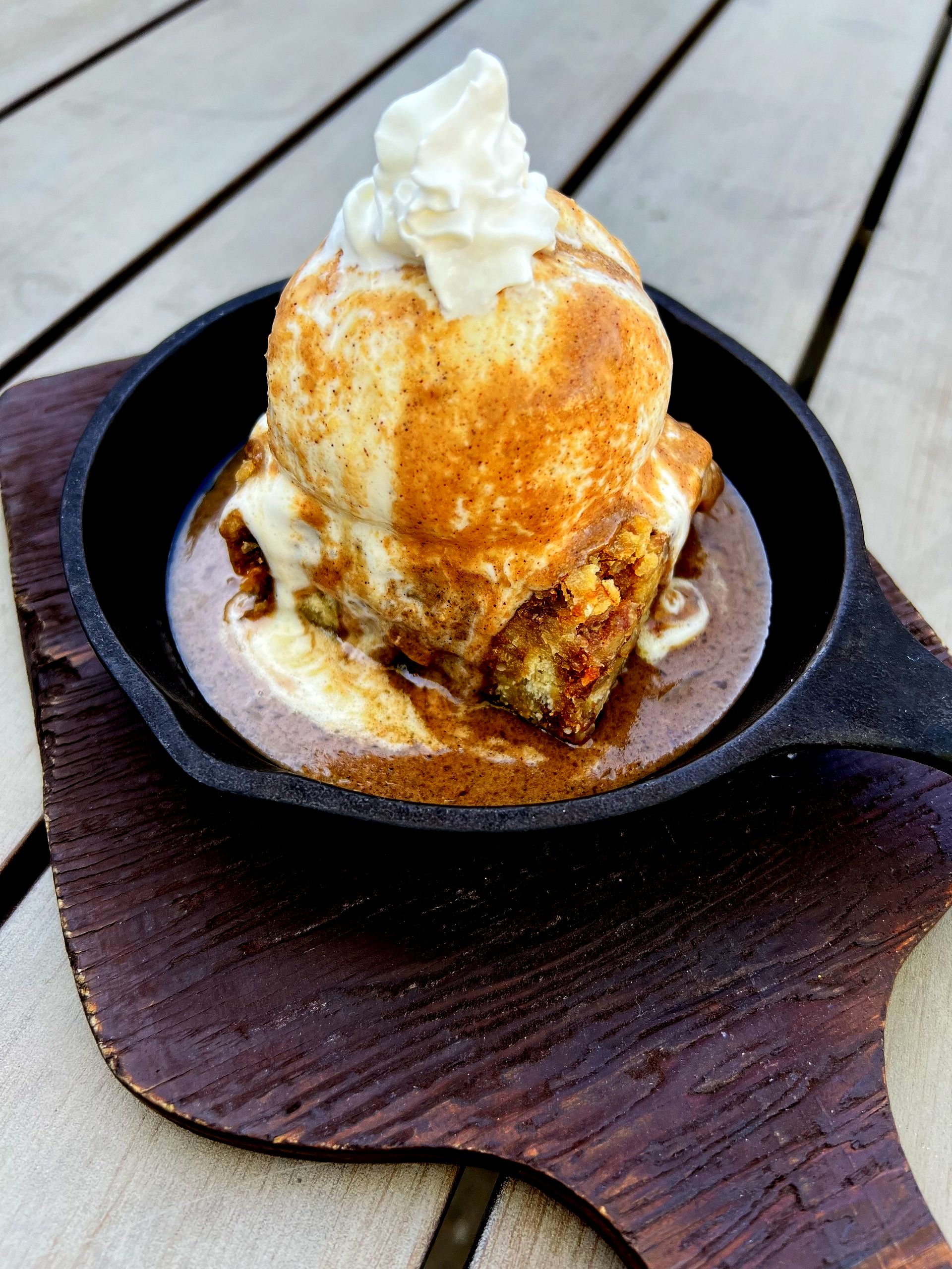 Skillet dessert: Bread pudding topped with ice cream, cinnamon, and whipped cream on a wooden board.