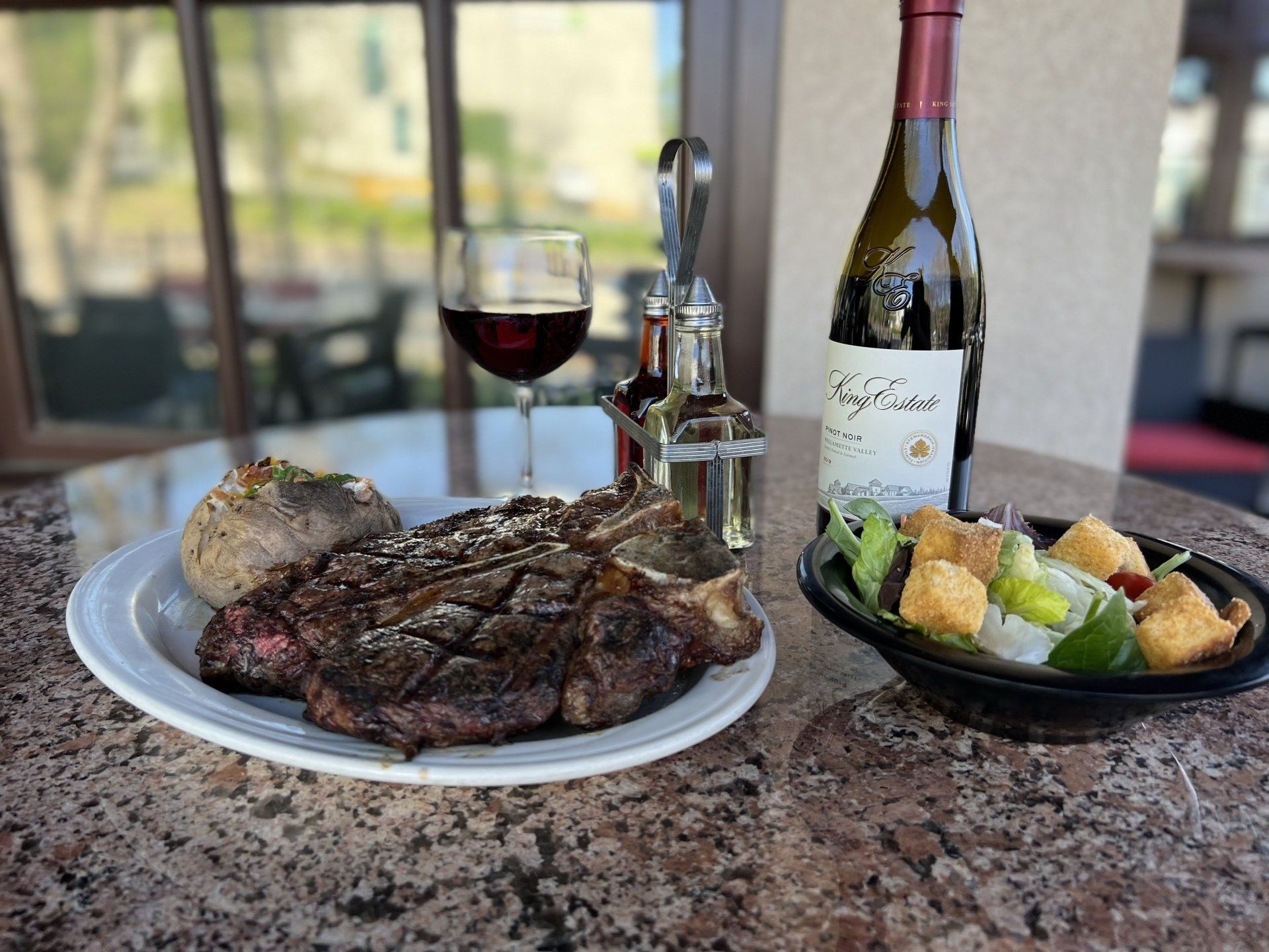 Steak dinner on a patio table with wine, a baked potato, and a salad.