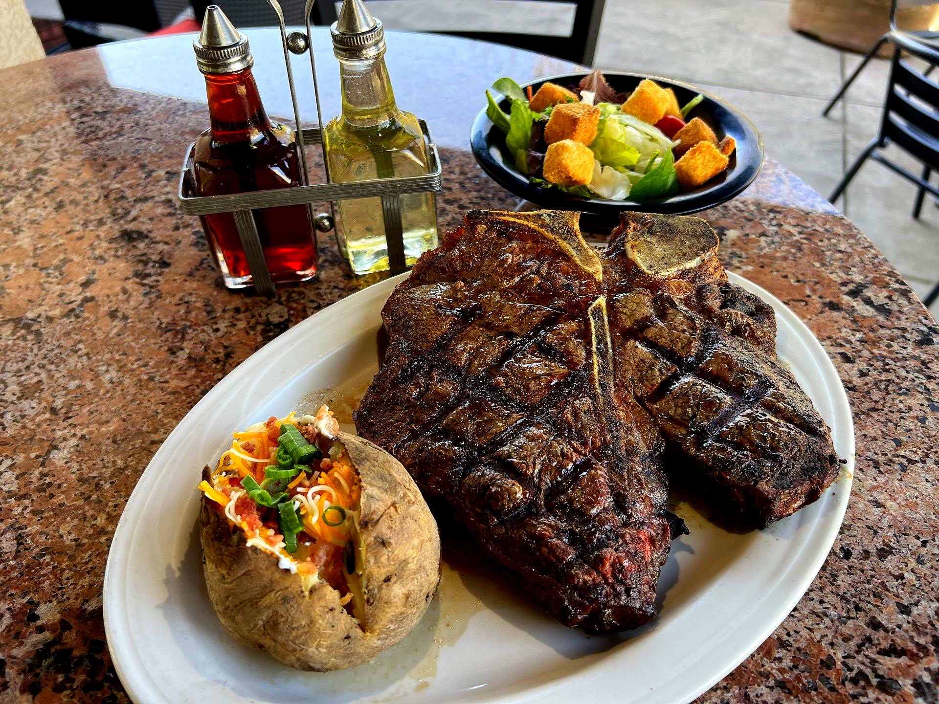 Grilled steak, loaded baked potato, salad, and oil bottles on a table.