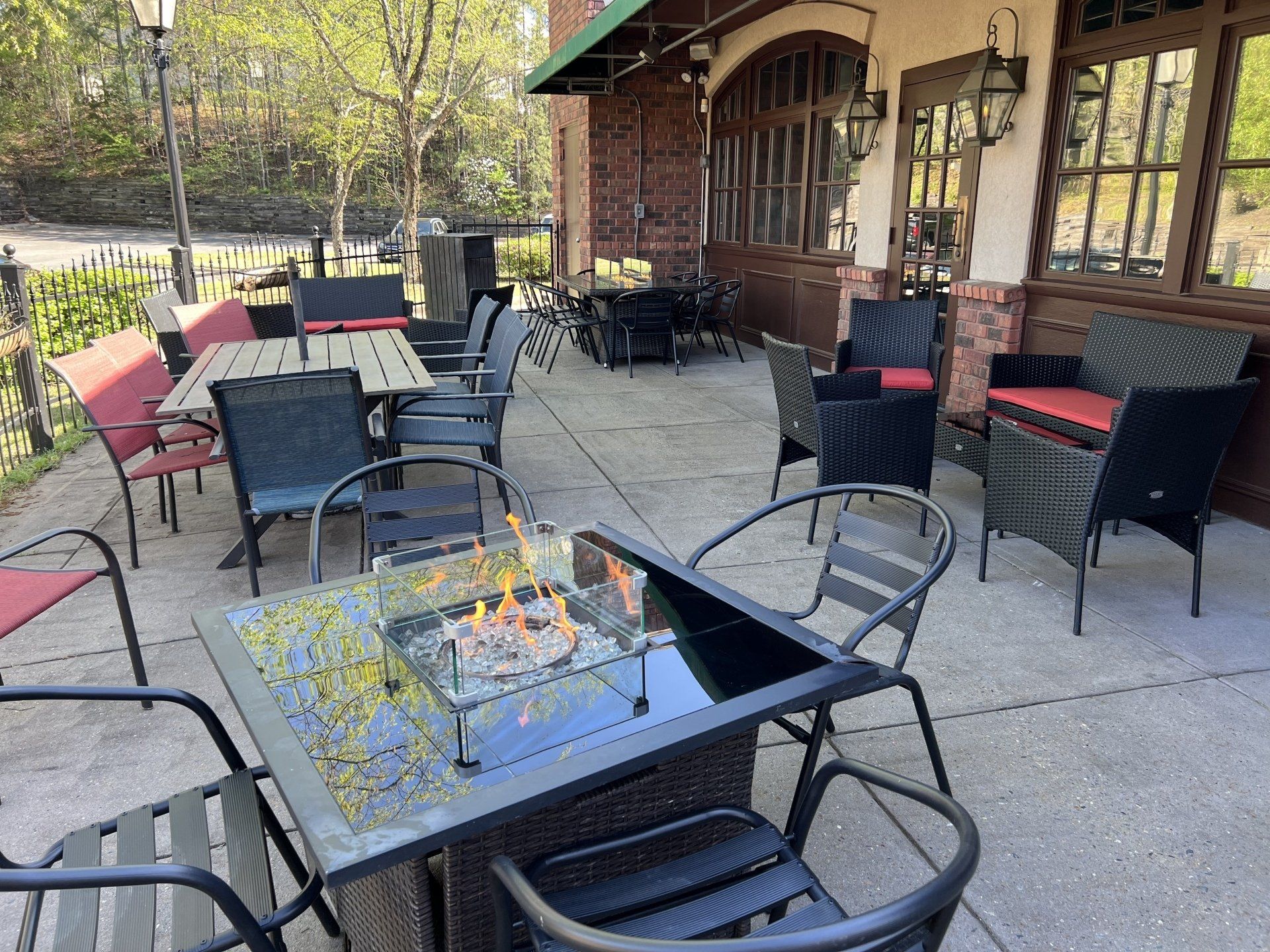 Outdoor restaurant seating area with tables, chairs, and a fire pit on a patio. Brick building background.