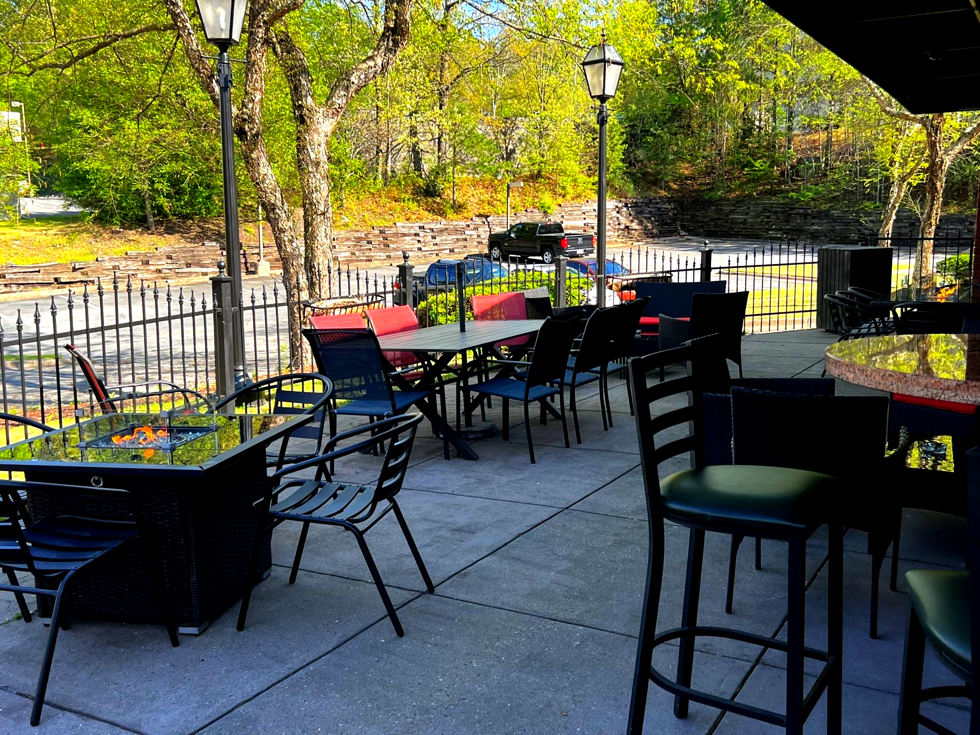 Outdoor patio with tables, chairs, and black metal fencing. Trees and greenery in the background.