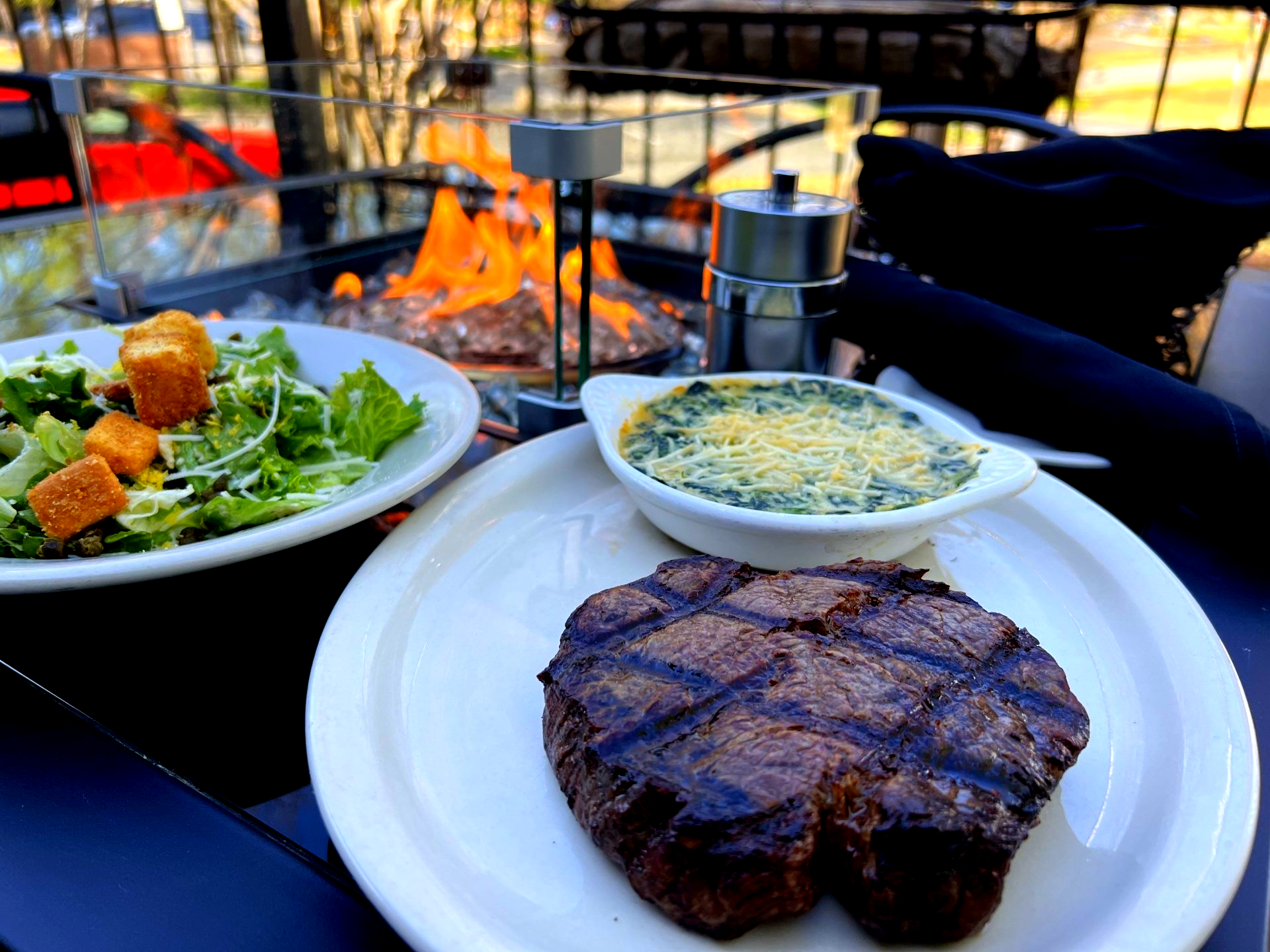 Grilled steak, salad, and spinach dip on a patio with a fire pit.