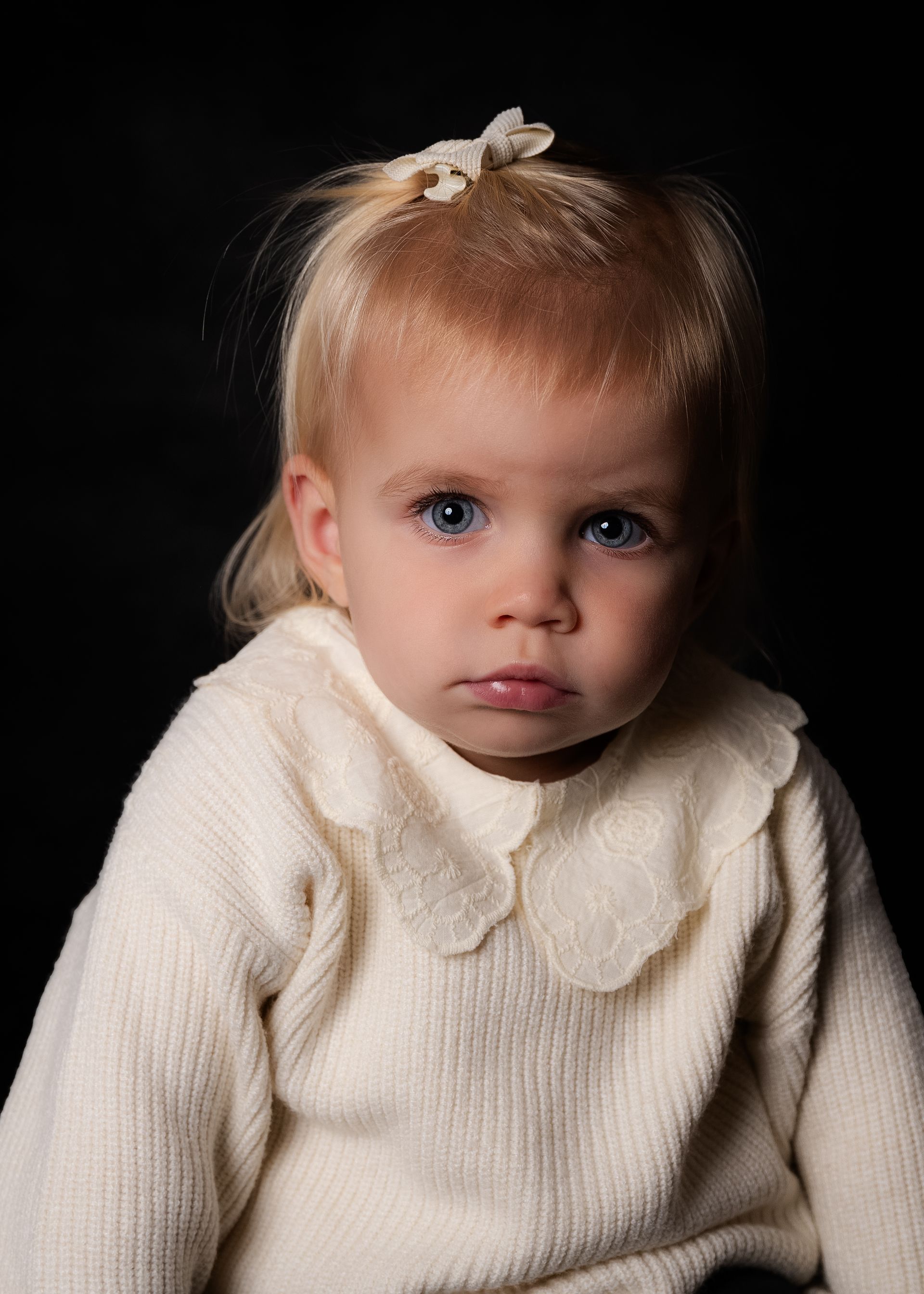 Een klein meisje draagt ​​een witte trui en een strik in haar haar.