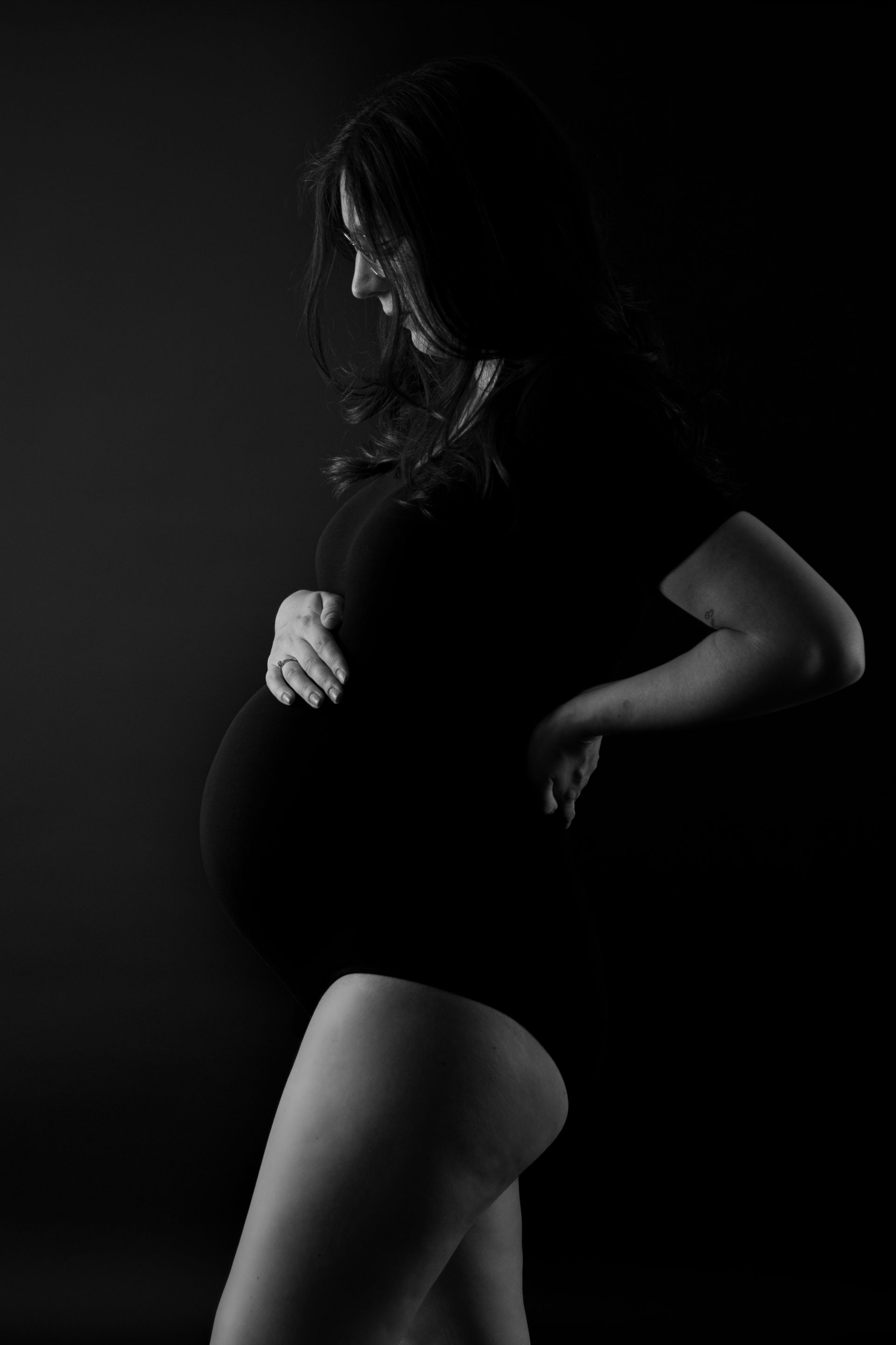 Een zwart-witfoto van een zwangere vrouw in een zwart bodysuit die haar buik vasthoudt.