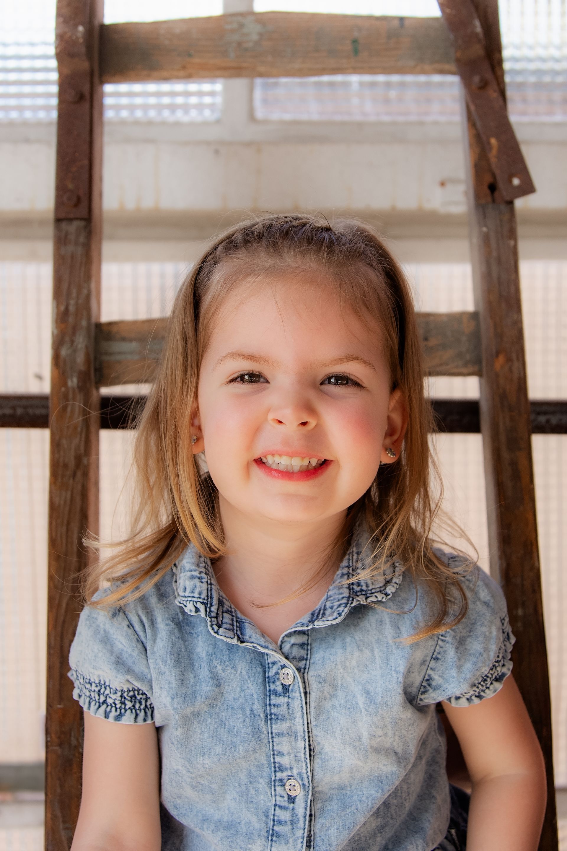 Een klein meisje zit op een houten ladder en lacht naar de camera.