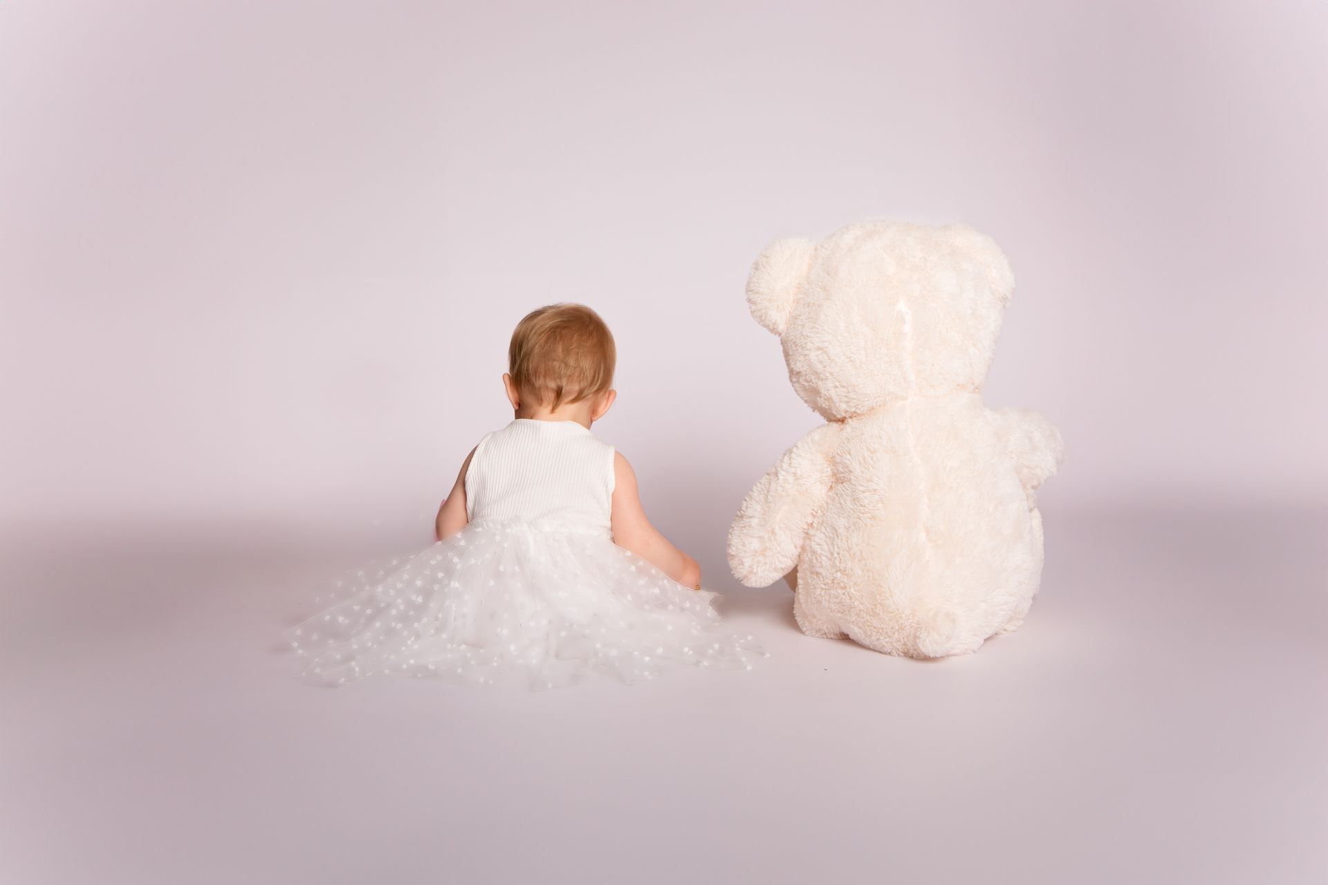 Een klein meisje in een wit jurkje zit naast een teddybeer.