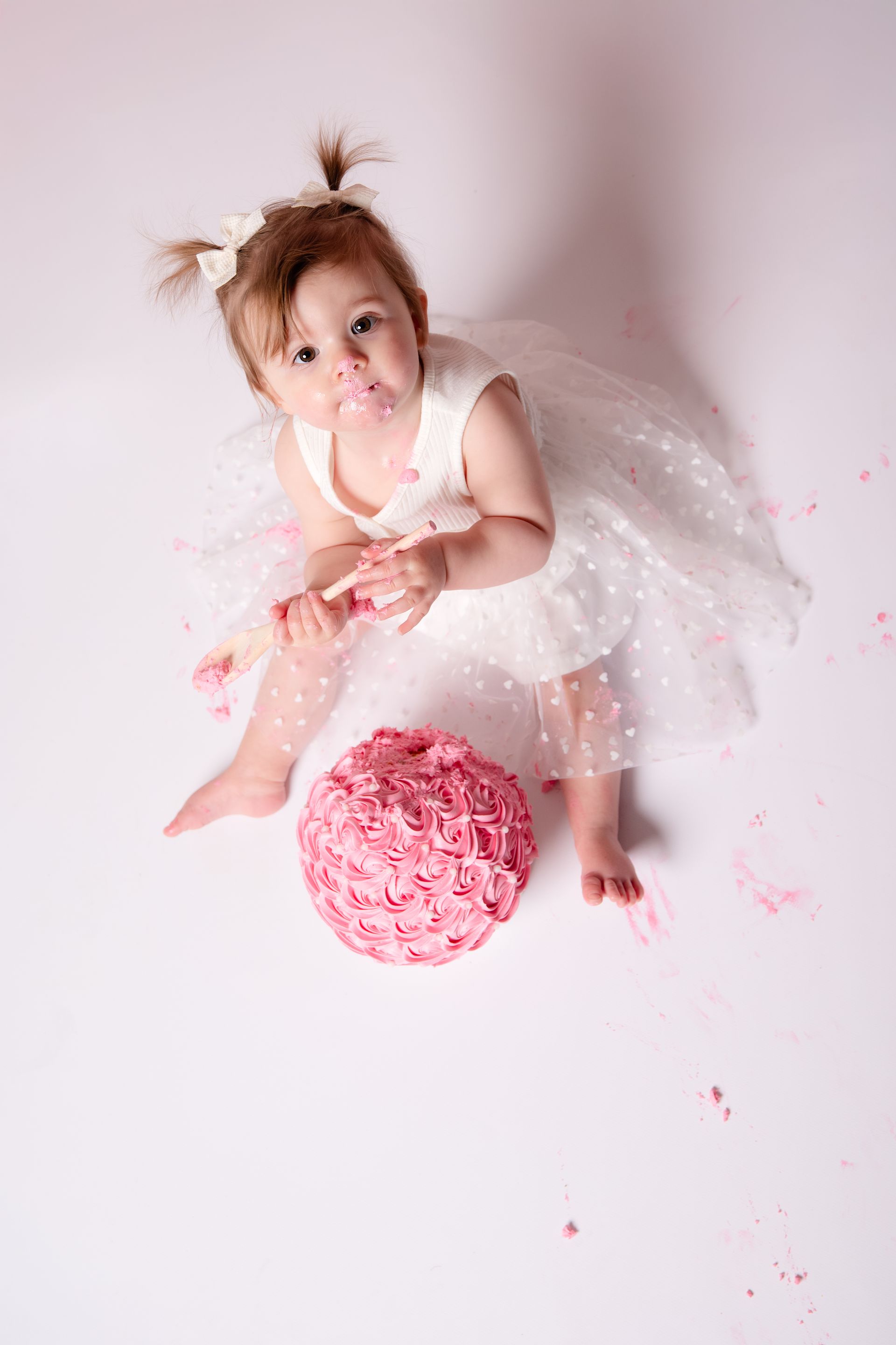 Een babymeisje in een wit jurkje zit naast een roze taart.