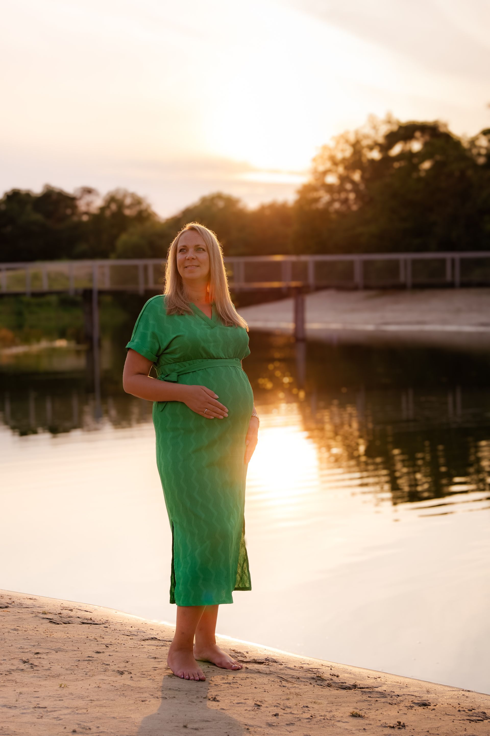 Een zwangere vrouw in een groene jurk staat op het strand bij een waterpartij.