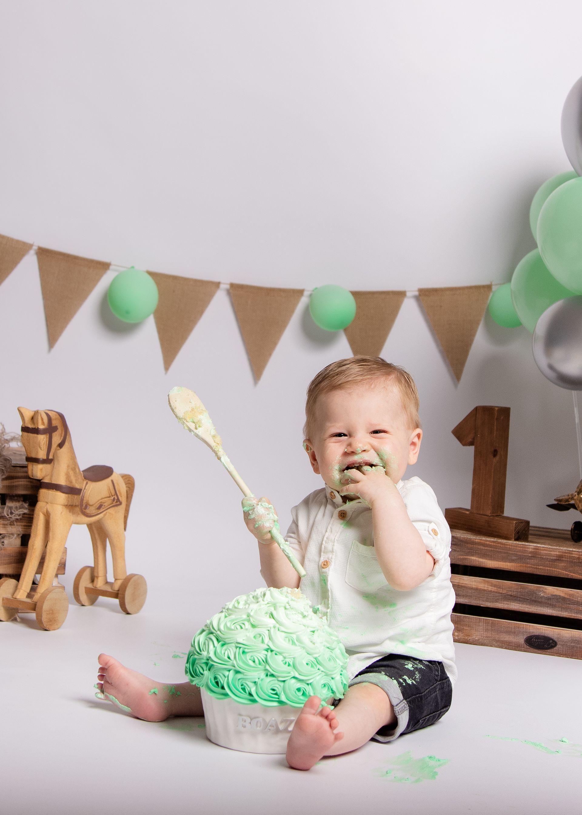 Een baby zit op de grond, eet een taart en houdt een lepel vast.
